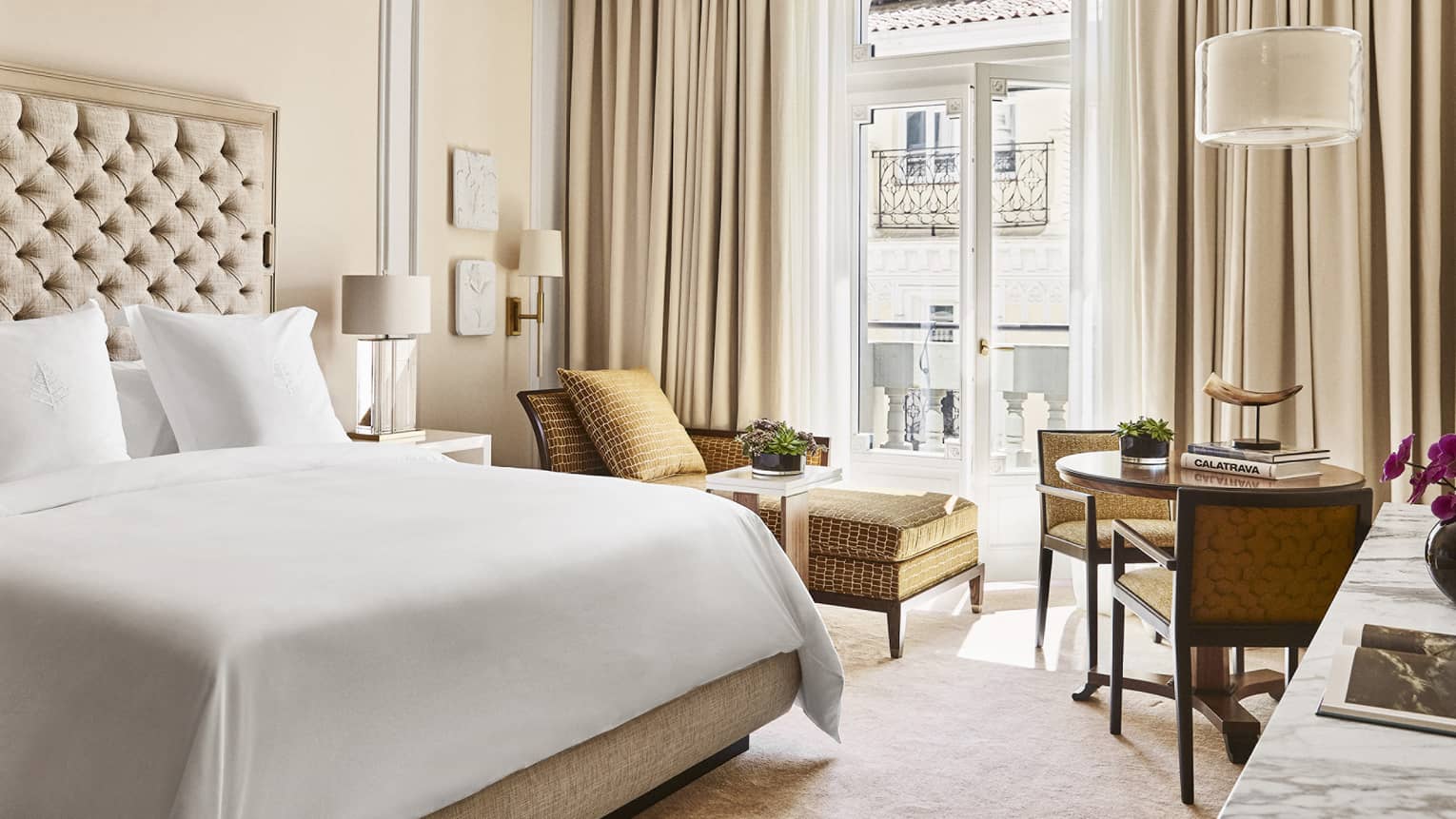 Guest room with king bed and white linens, tan chaise lounge, two-seater table, French doors opening onto balcony