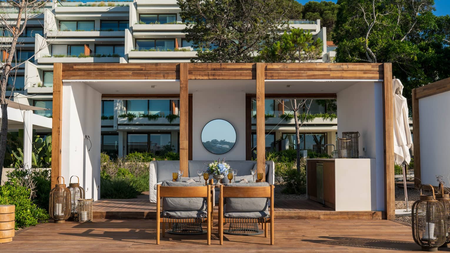 A pristine white cabana with wood accents frames a patio dining area in front of hotel balconies laden with greenery. 
