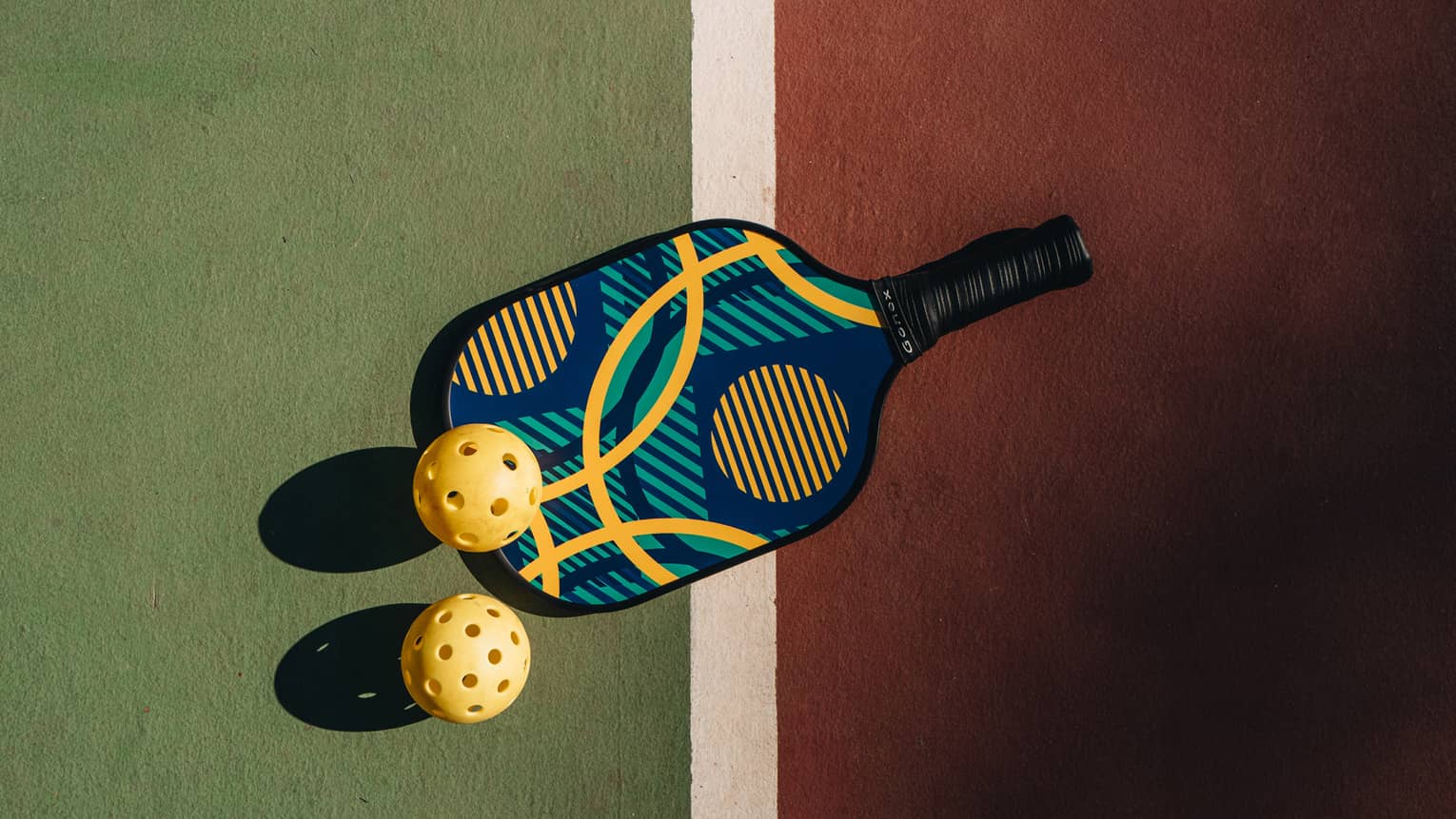 A colourful pickleball racquet and two yellow pickleballs cast shadows upon a green and red court with a white dividing line.