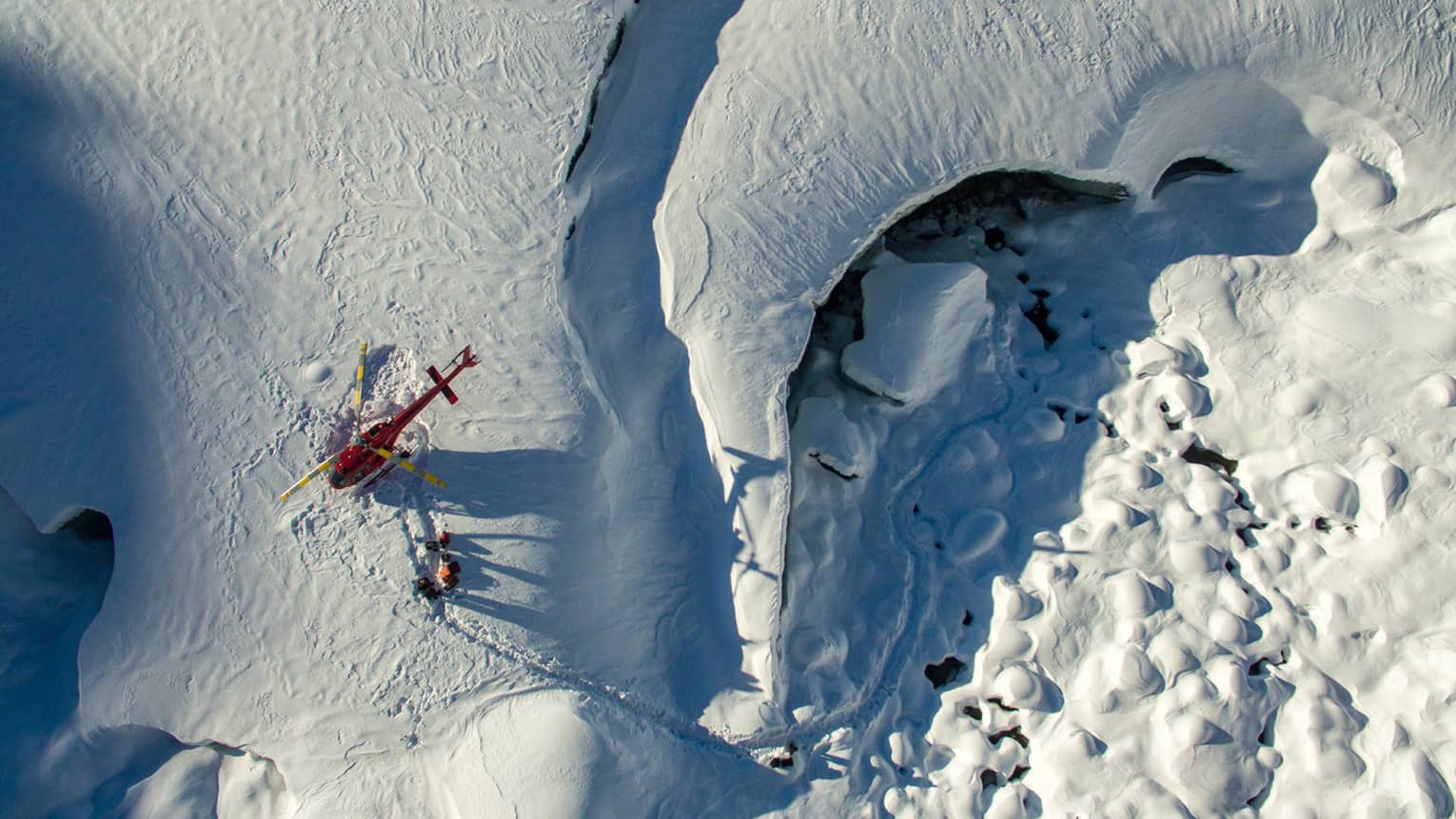 Aerial view of a red helicopter that's landed on a snow-covered mountain with three people walking near it