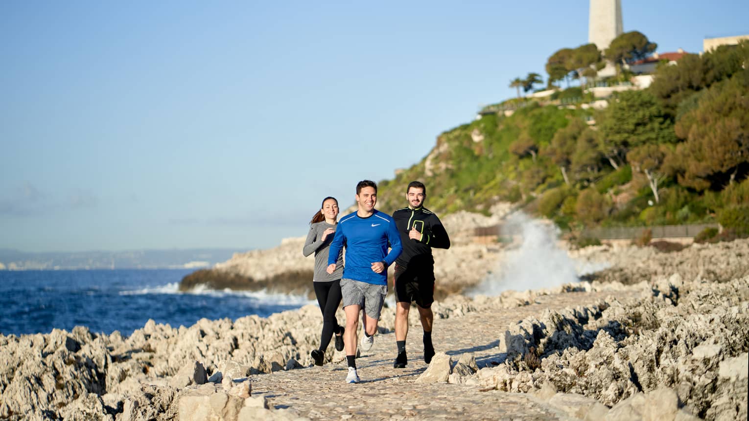 Three people jogging, smiling, on rocky trail beside the sea, lighthouse in backdrop