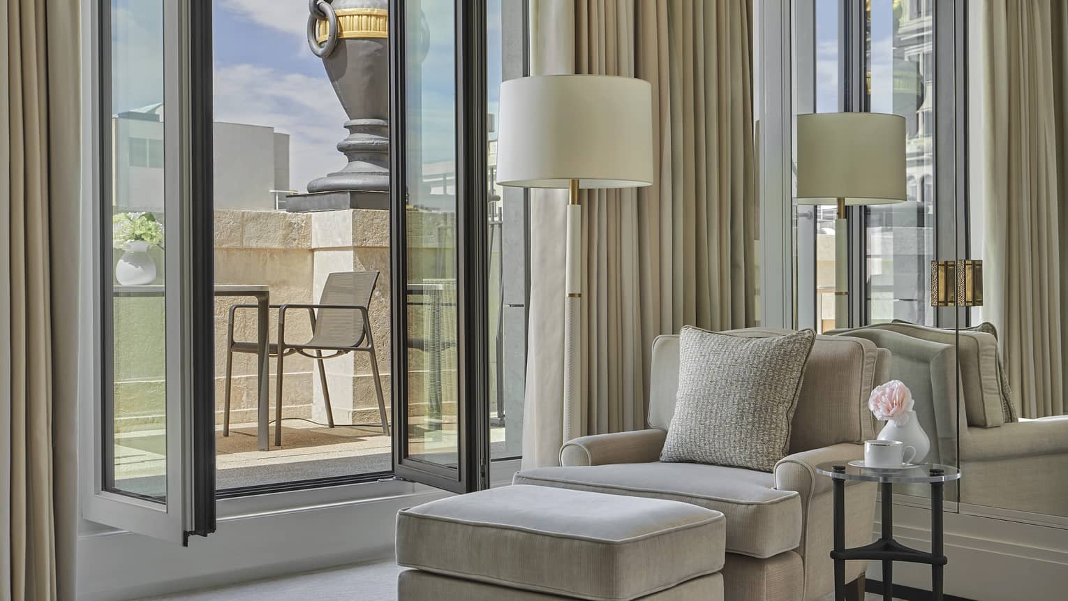 Guest suite with cream accent chair and matching ottoman, floor lamp, floor-to-ceiling window with cream curtains, glass door opening onto terrace