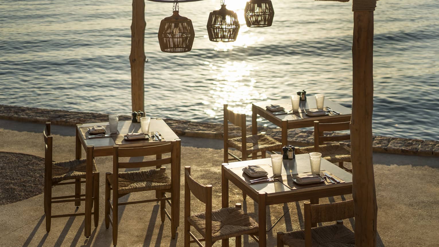 Outdoor taverna next to ocean with three wooden tables and chairs, three wooden lamps