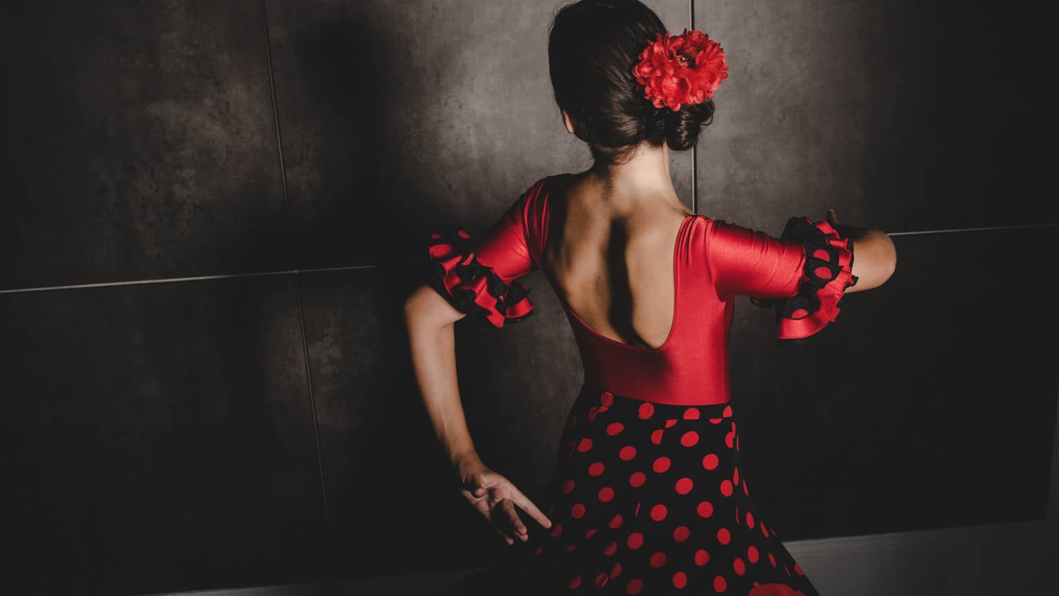 A woman wearing a black and red flamenco dress and red flower in her hair dances with her face turned away from the camera 