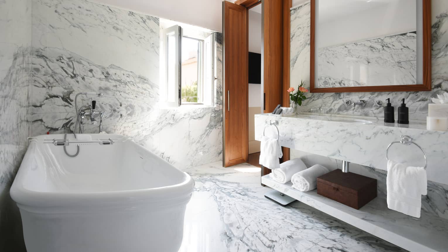 A bathroom with marble walls and floor, a tub, a. large mirror, and bath accessories.