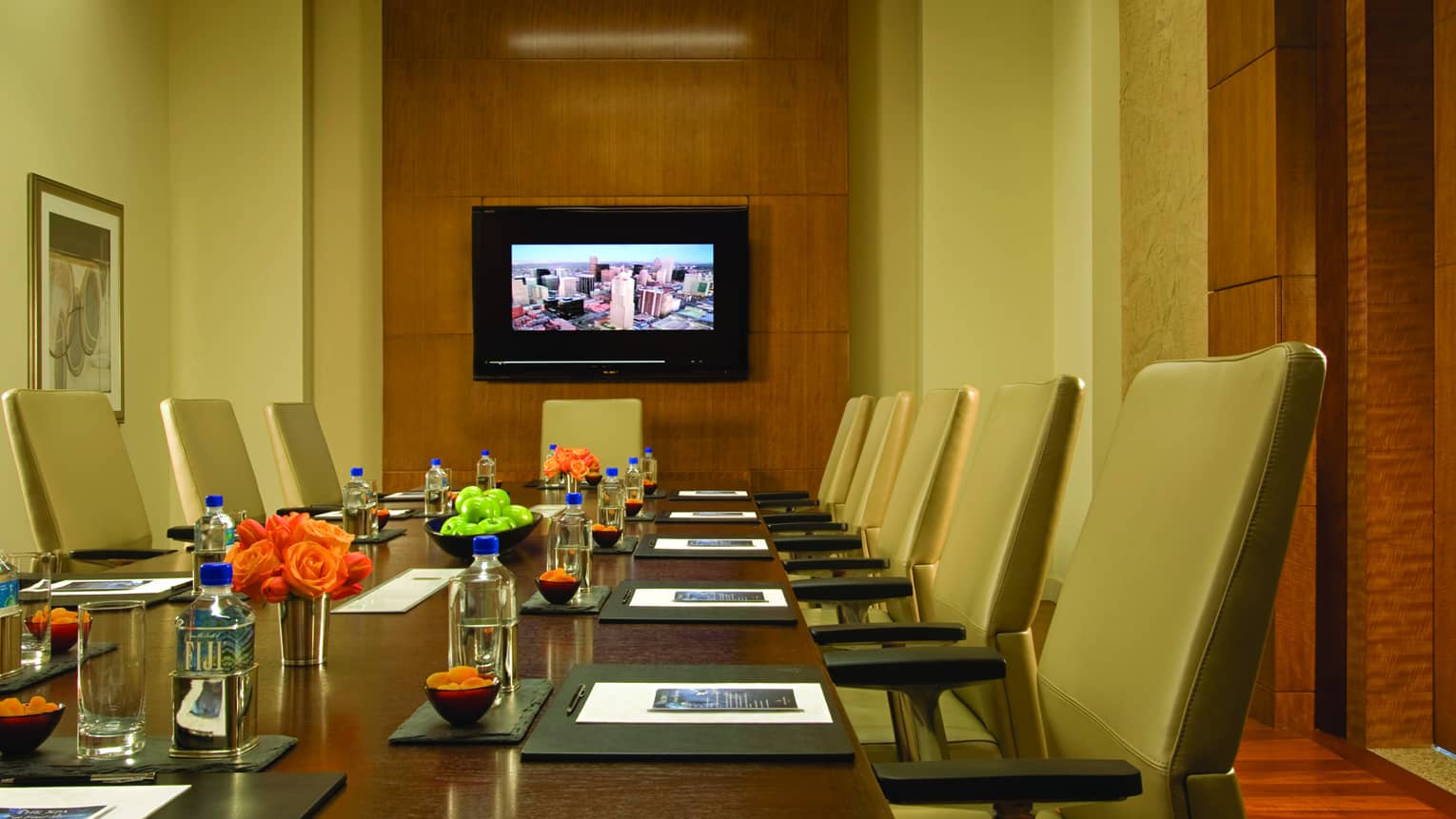 Executive boardroom table with tan leather chairs, flowers by modern wood panel wall with TV screen