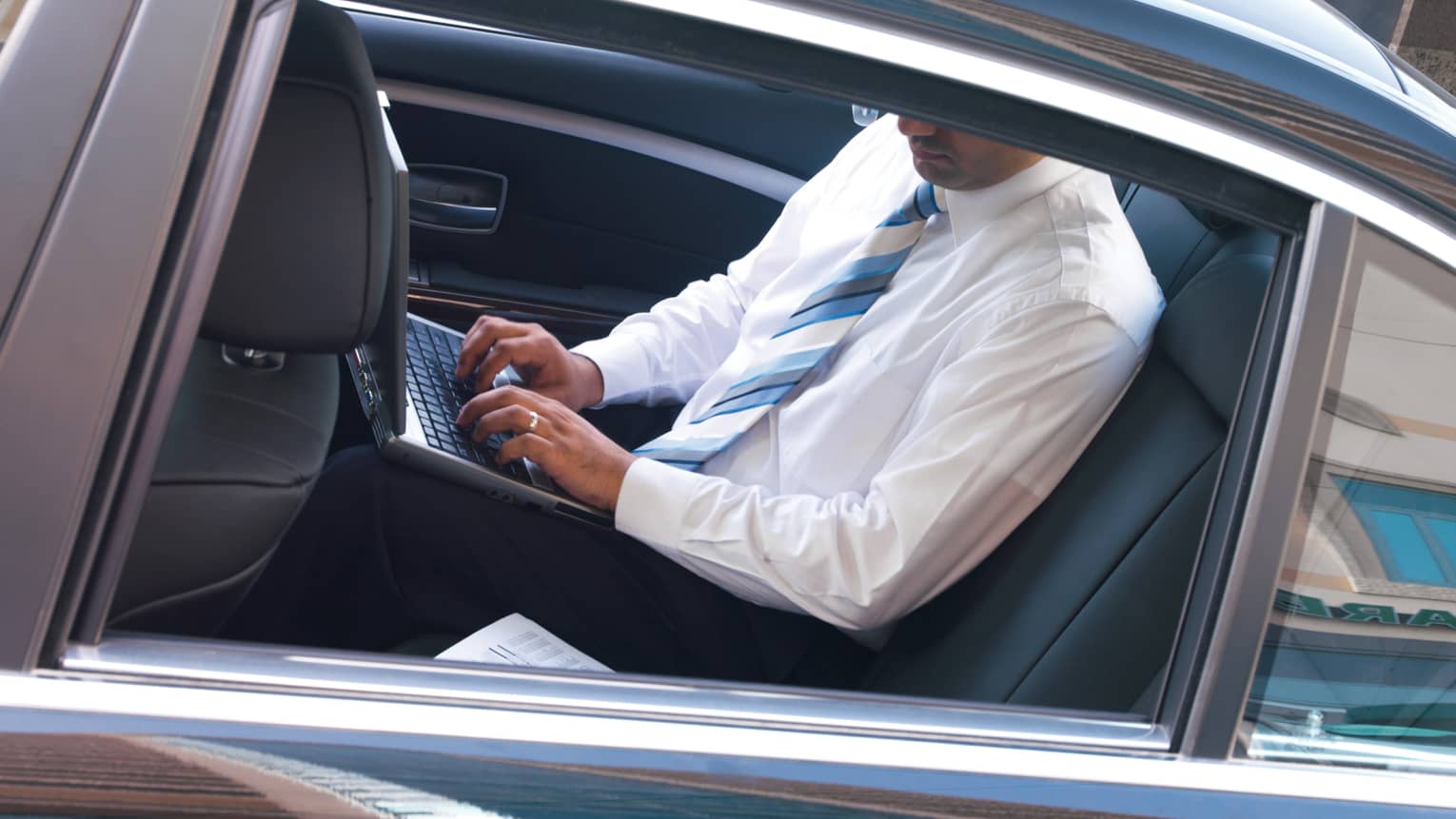 View of car window where man in white dress shirt and blue-and-white tie sits in backseat, works on laptop computer