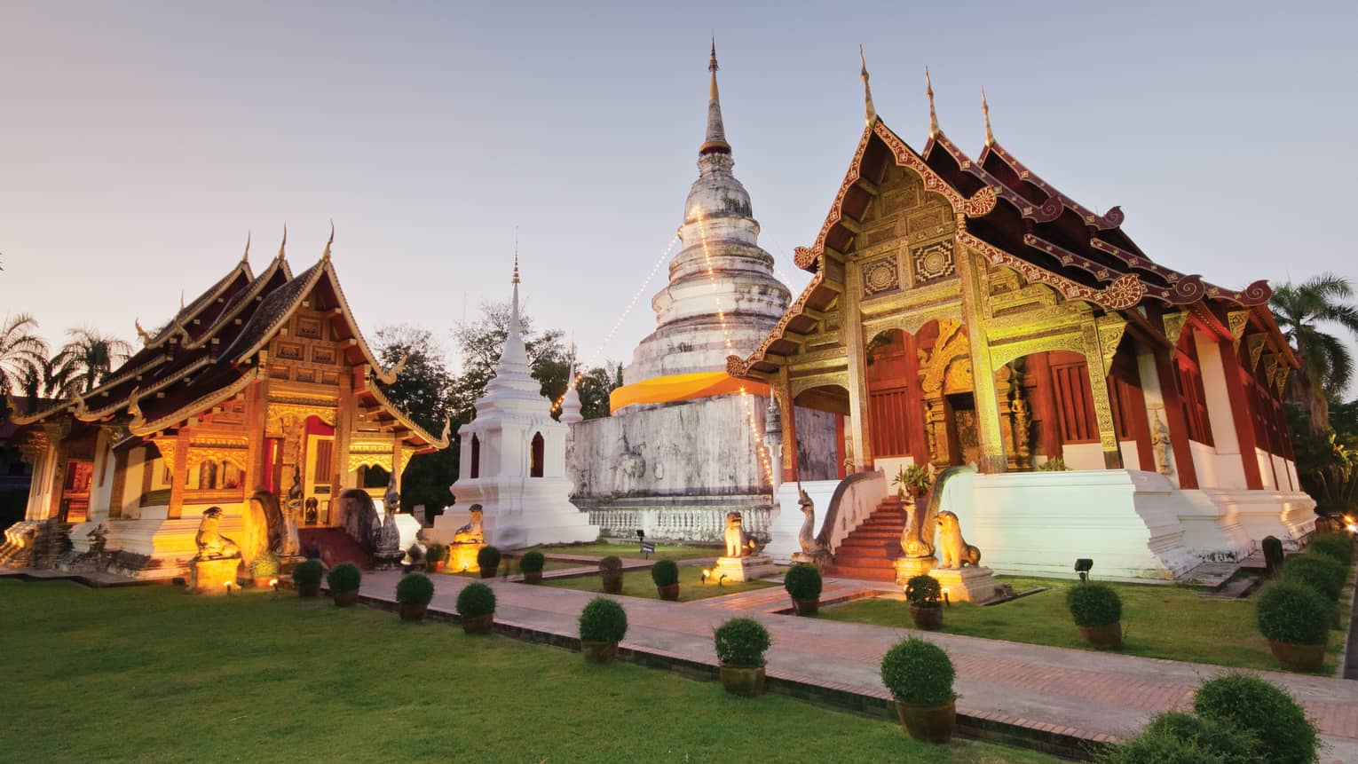 Two temples with gabled roofs and intricate carvings uplit in gold, between them a white domed building and stone monument.