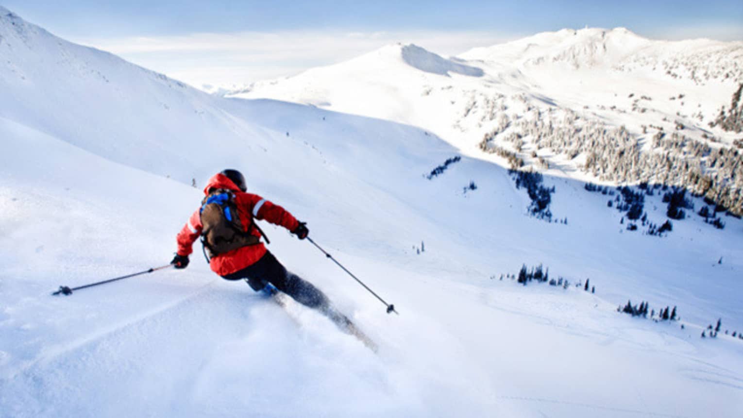 Back view of skier in red jacket, backpack, skiing down mountain slope on sunny day