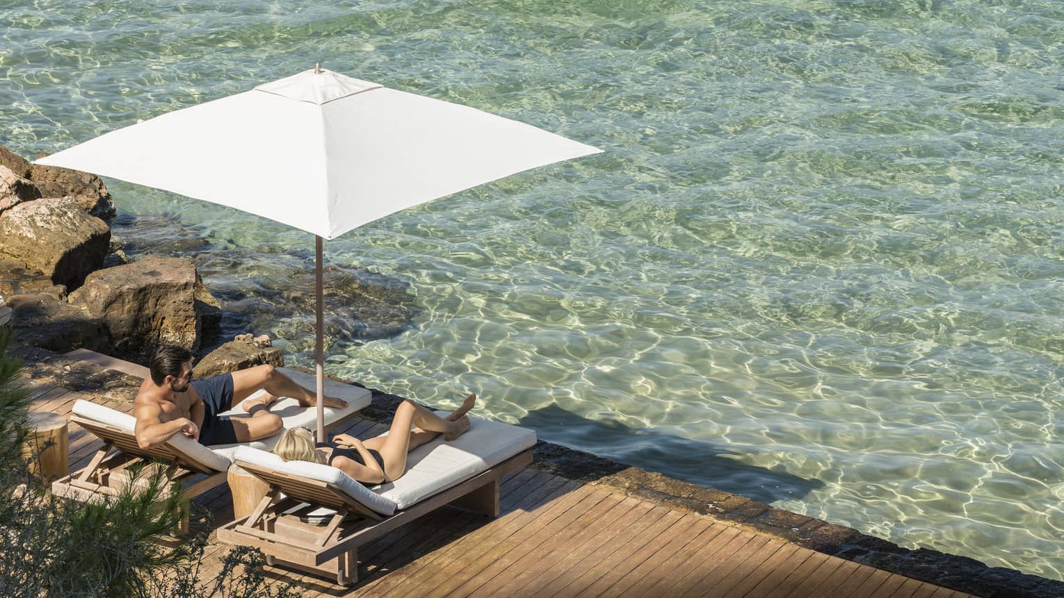Couple lounges on deck chairs under umbrella on oceanside patio