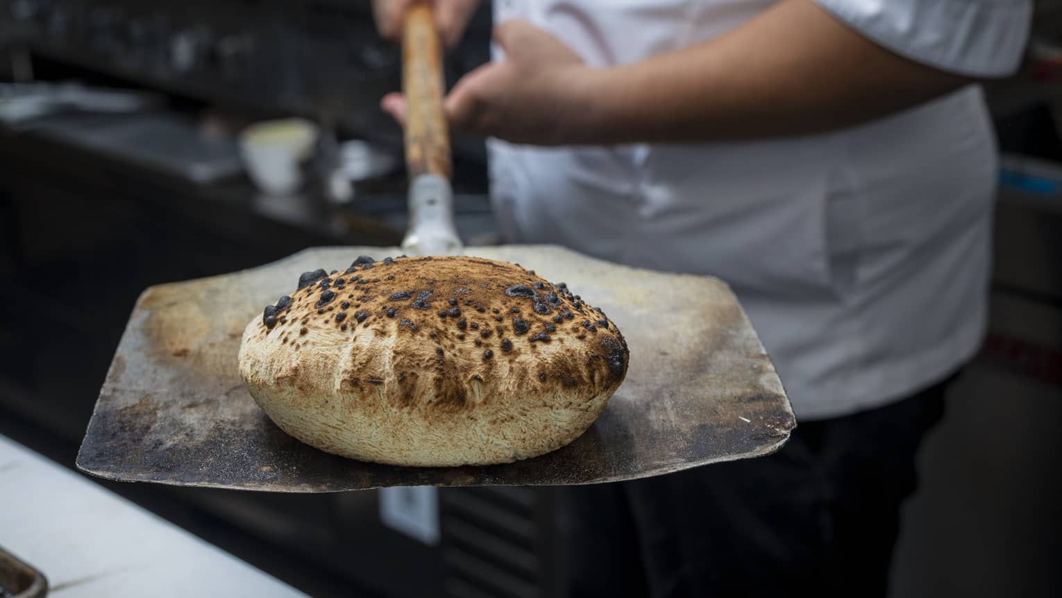 A chef wearing a white chef's jacket holds a fresh-baked loaf of bread on a large peel