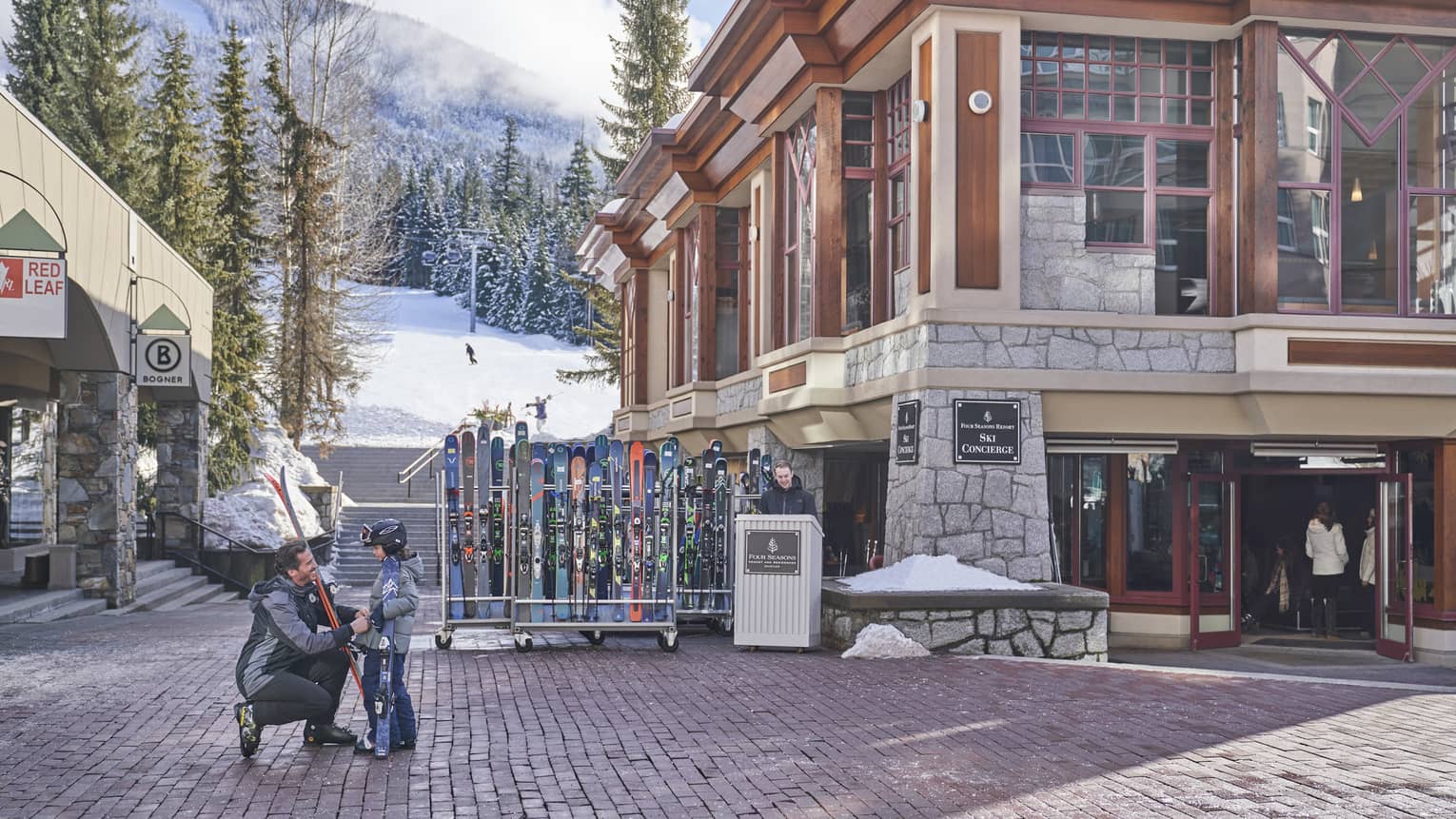 Man kneeling next to child, with skies, outside in front of ski shop, mountain in background