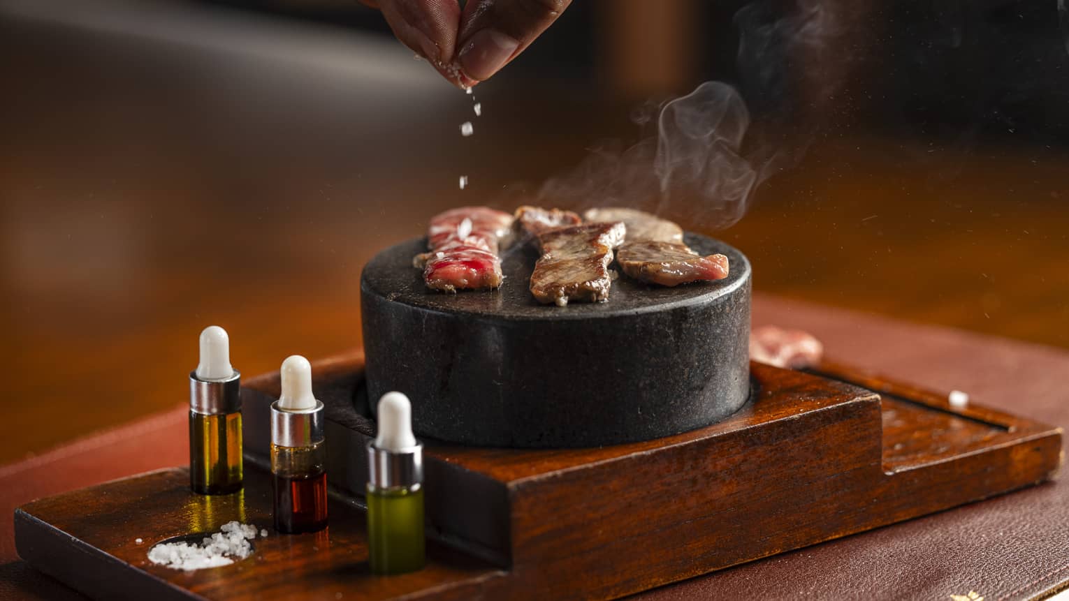 A hand sprinkles salt on small pieces of beef as they cook on a small, black table-top barbecue grill