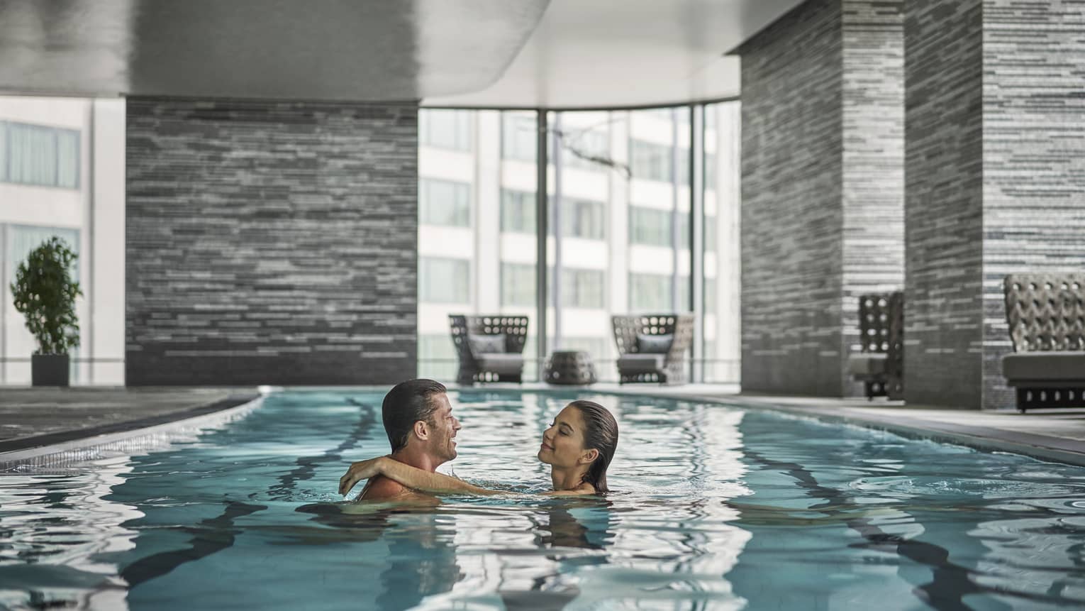 A couple embraces in a lap pool; behind them, floor-to-ceiling windows reveal an urban landscape.