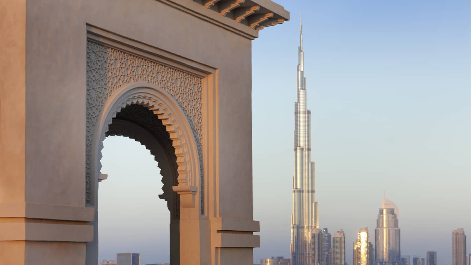 Orange glow of sunset on a decorative stone arch, the iconic Burj Khalifa gleaming in the distance against blue sky.,Orange glow of sunset on a decorative stone arch, the iconic Burj Khalifa gleaming in the distance against blue sky.