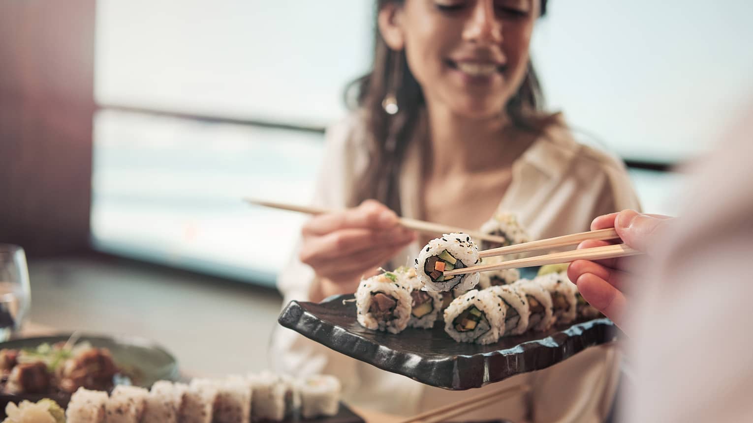 A woman picks up a sushi roll with a set of chopsticks off of a tray of sushi.