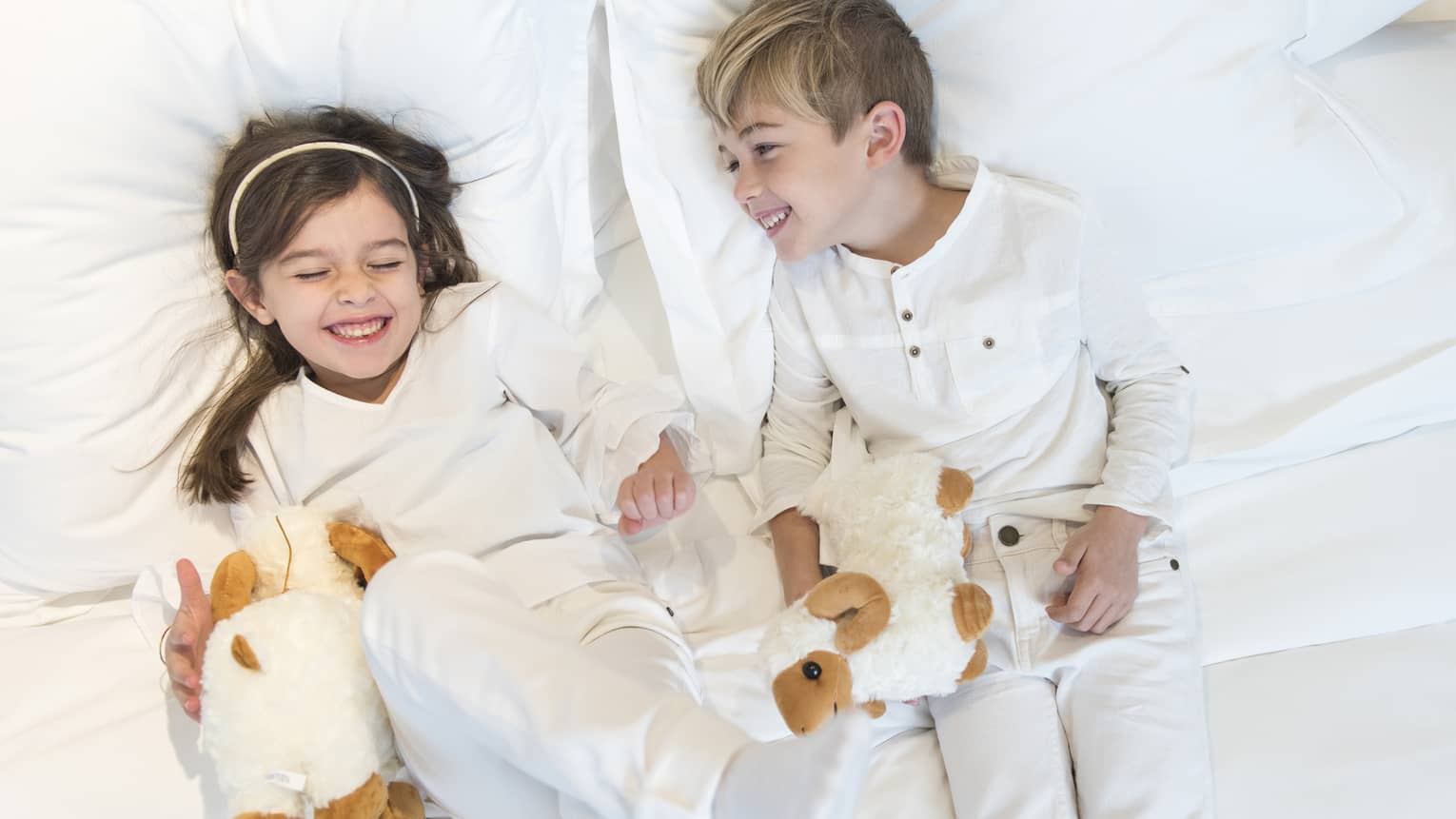 Aerial view of two children wearing white pyjamas with white stuffed animals lying on bed, laughing