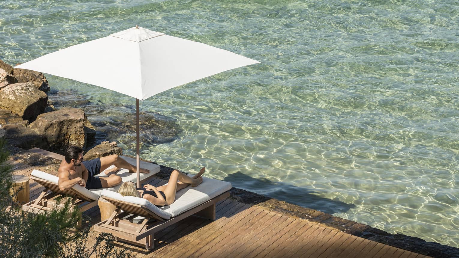 Couple lounges on deck chairs under umbrella on oceanside patio