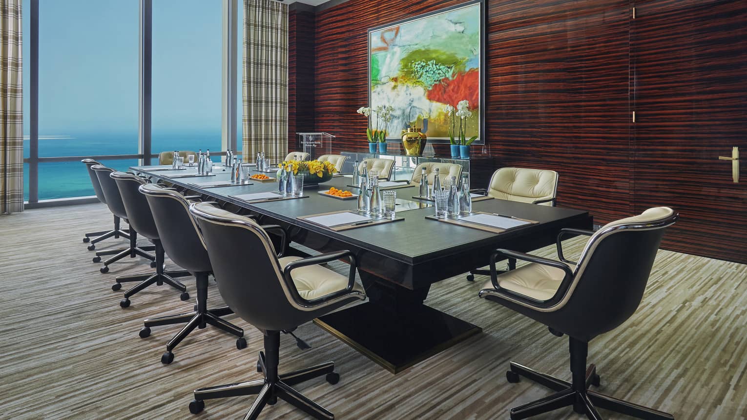 Boardroom meeting table lined with swivel chairs by wood wall with large painting, window
