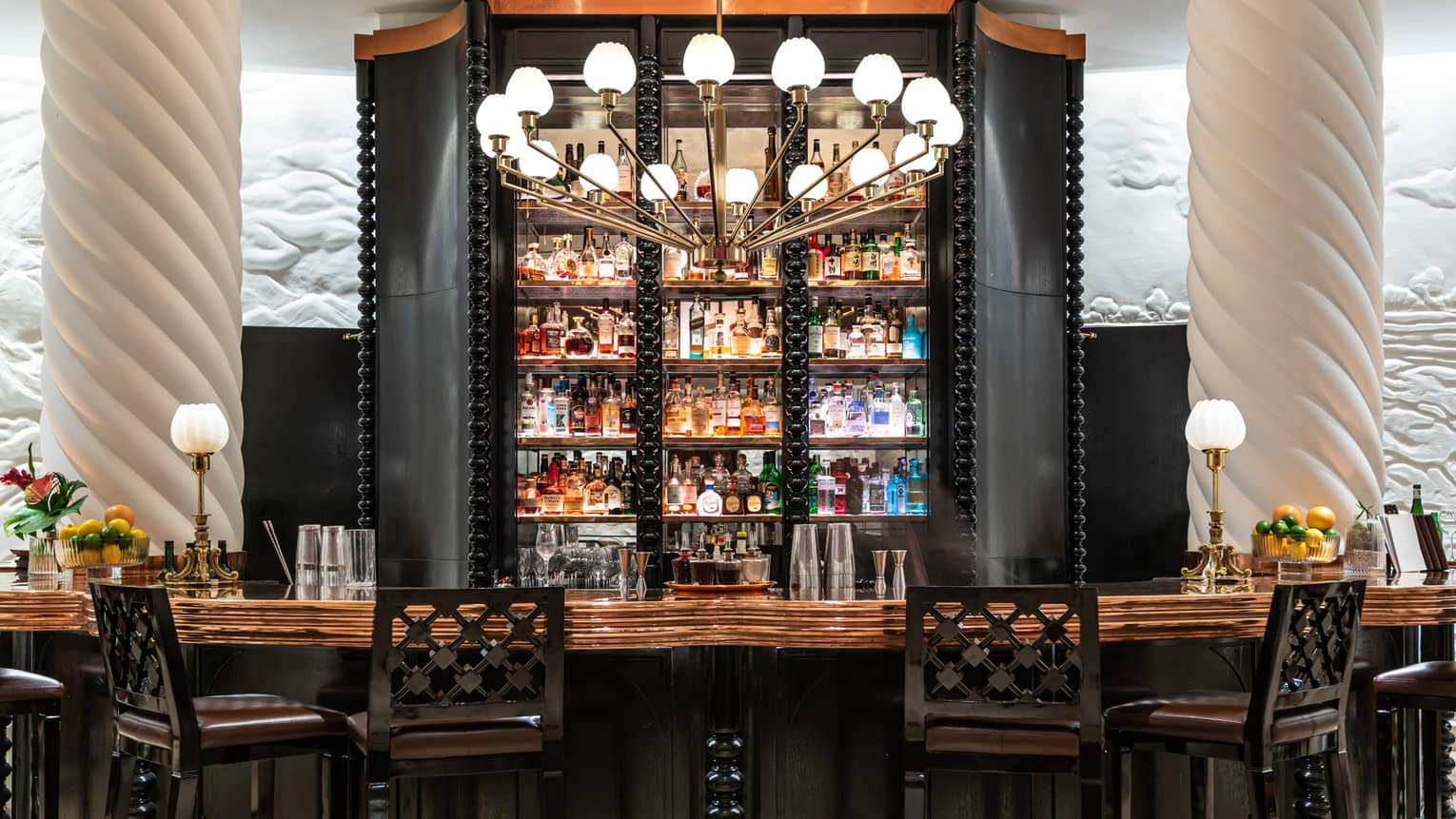 Wall of liquor bottles behind the wooden bar, four black barstools in front, white columns on either side