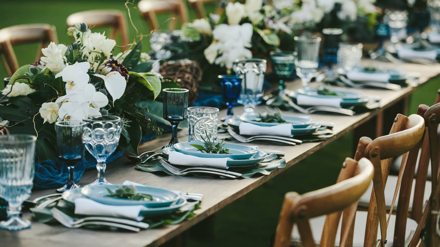 A close up of a long wooden table set with blue plates, eclectic blue and clear glassware, white napkins and green and white floral arrangements all the way down the center of the table