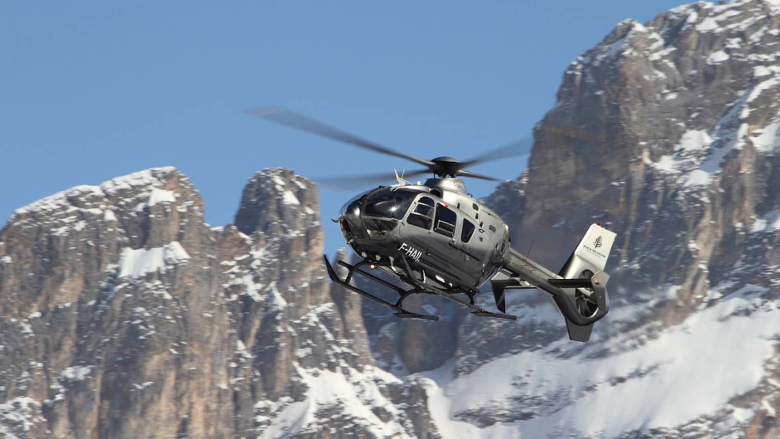 Silver helicopter ascending beside snowy, rocky mountain peaks