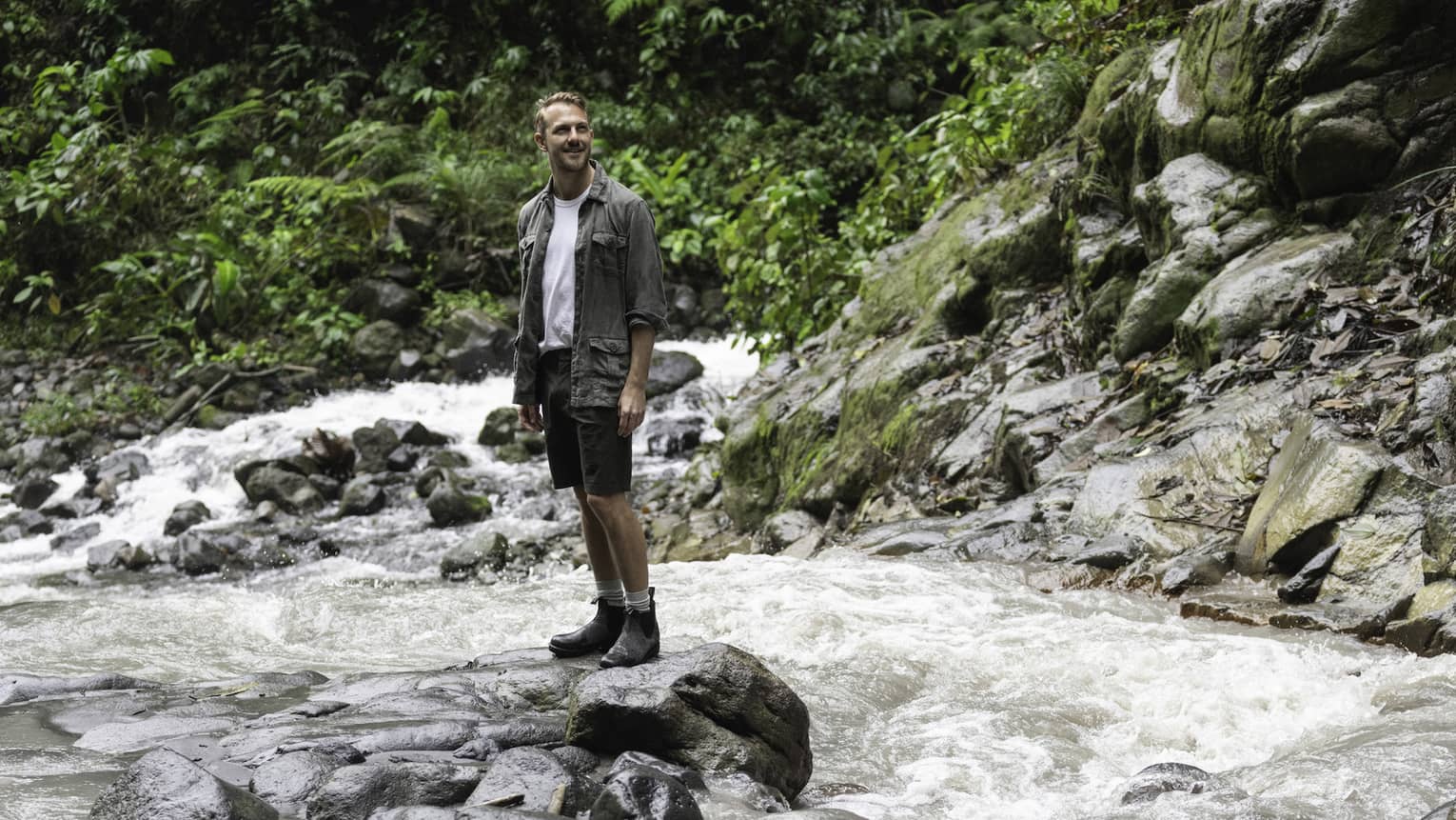 Man wearing hiking jacket, white t-shirt, shorts and hiking boots stands on a cluster of rocks in the middle of a rushing river