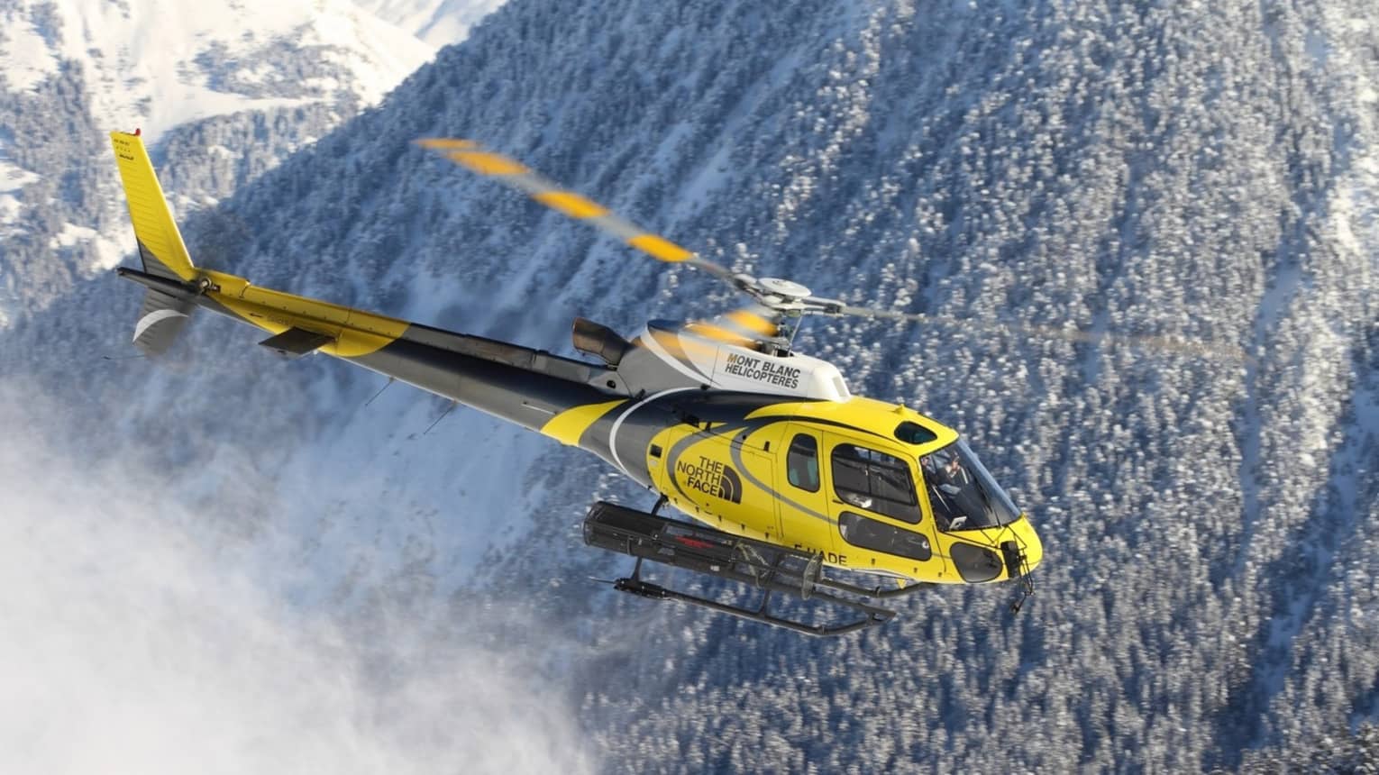 A yellow helicopter flies through a snow covered mountain in the swiss alps