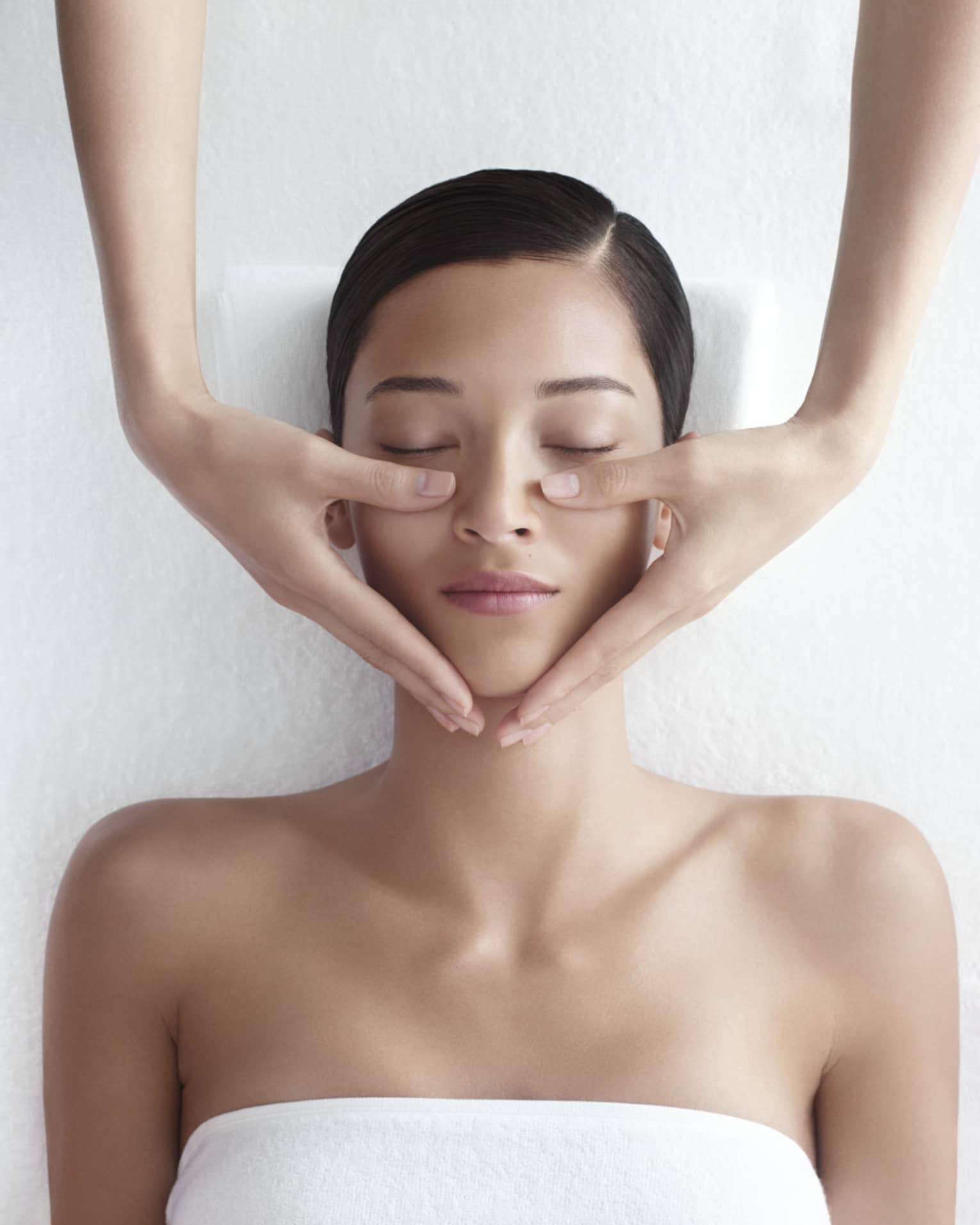 Woman in white bathrobe lies on massage table with eyes closed, masseuse rests hands on her cheeks