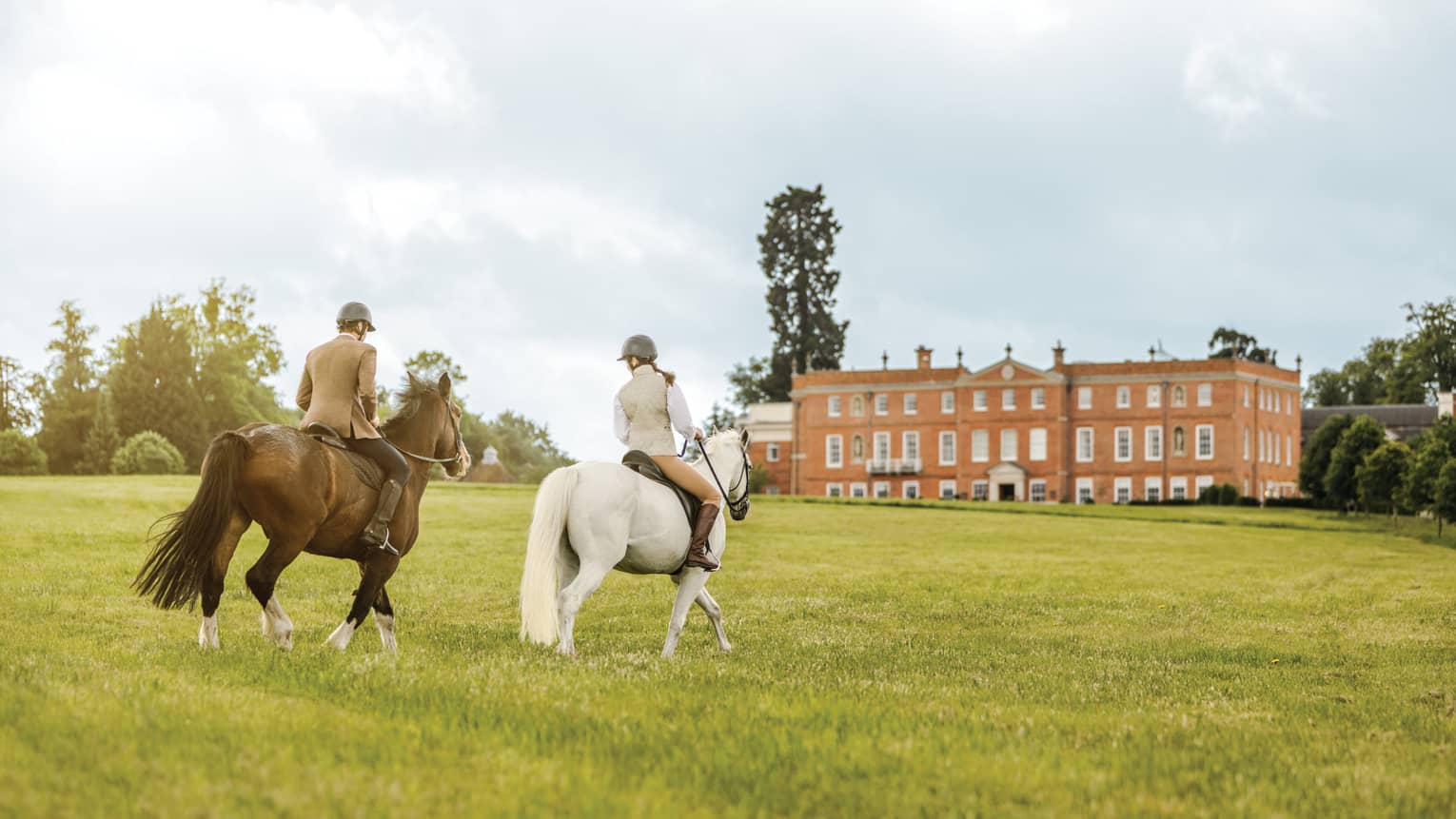 Couple rides horses across green English field to Four Seasons Hotel Hampshire building