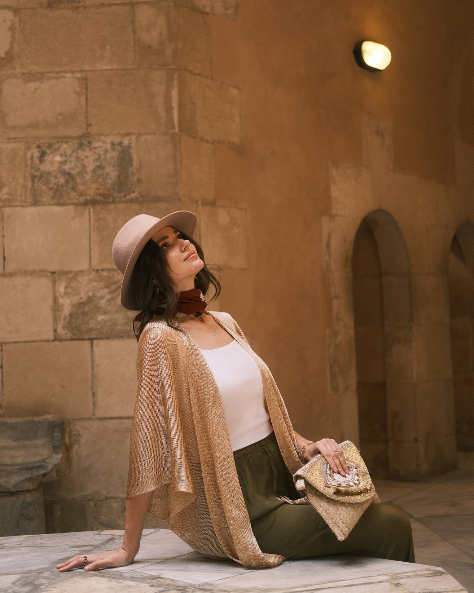 A modestly dressed guest reclines on a stone bench gazing upward, behind them stone walls and arched doorways.