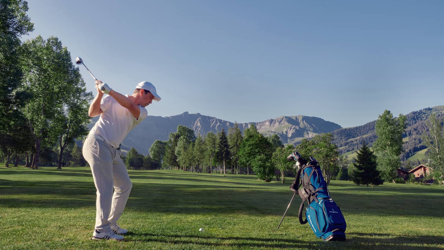 Man on mountainside golf course swings to hit golf ball beside blue golf bag