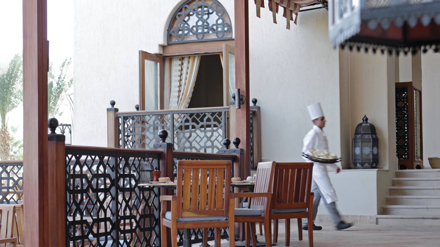 Chef in white uniform walks past wood patio tables, chairs, decorative railing at Arabesque restaurant