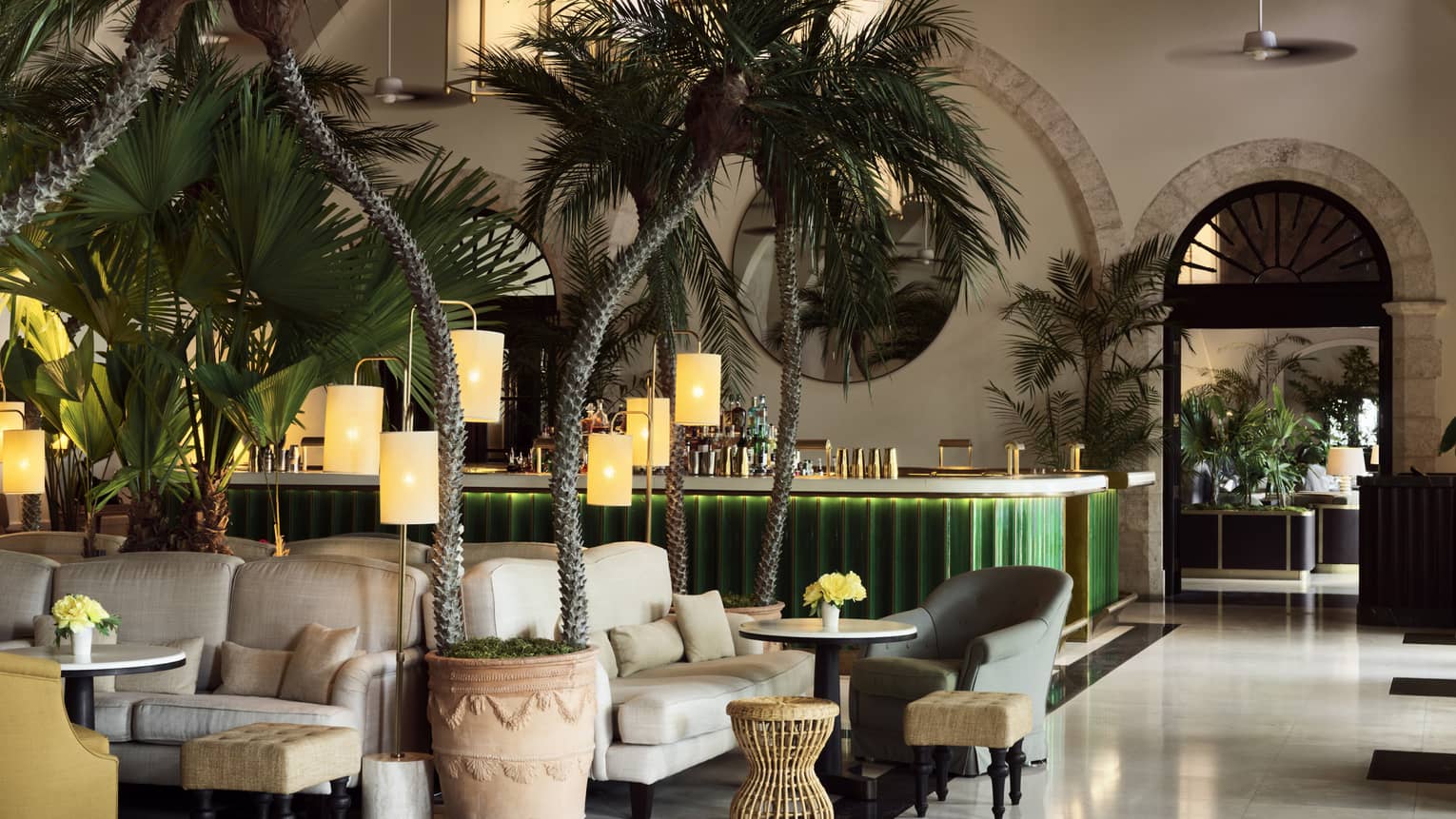 Indoor lounge at the Champagne Bar, with palm trees, white upholstered arm chairs and a green bar in the background. One of the many Surfside restaurants of note at Four Seasons Surf Club, Florida.