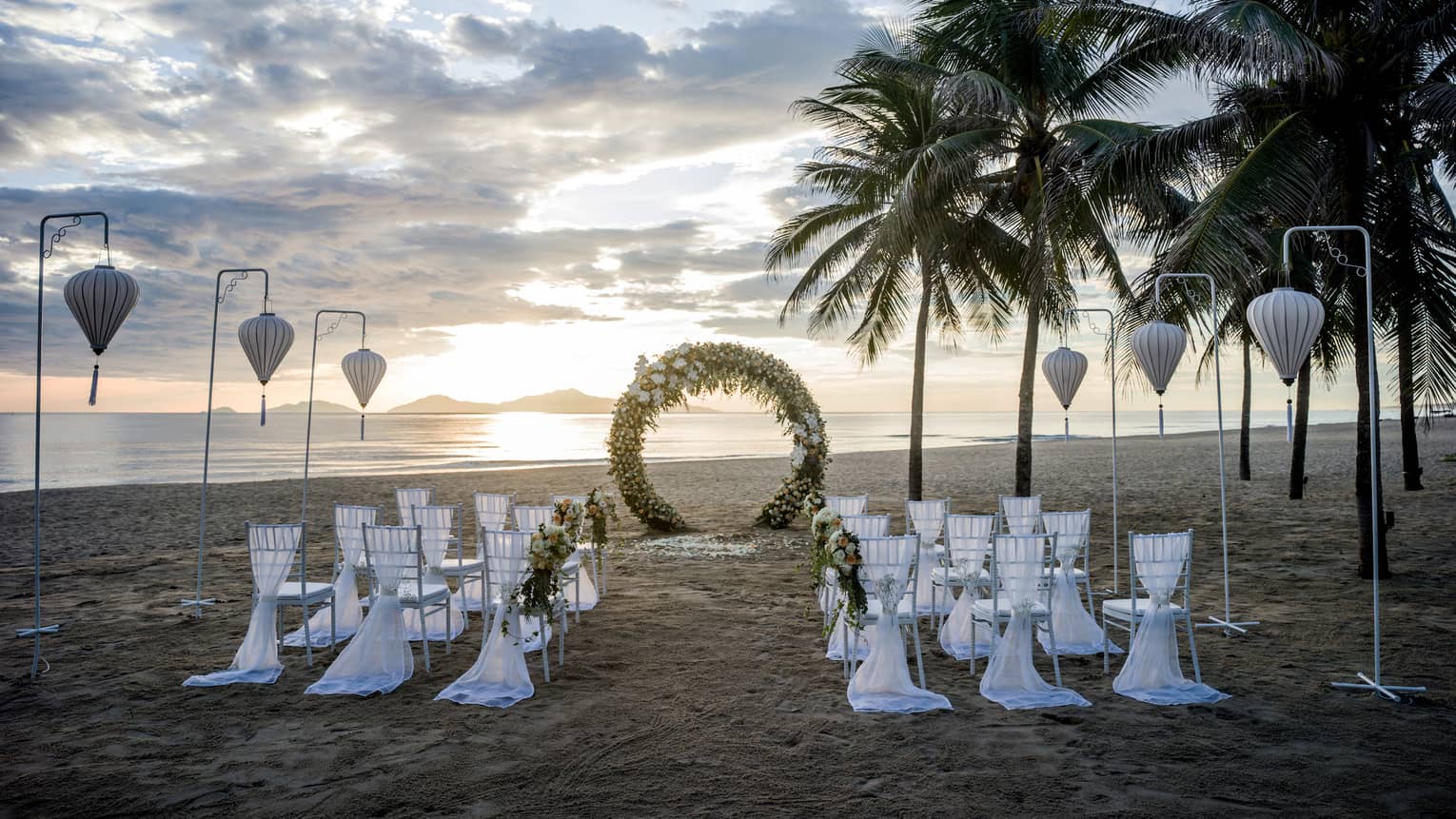 Rows of white chairs face round wedding altar on sand beach at sunset