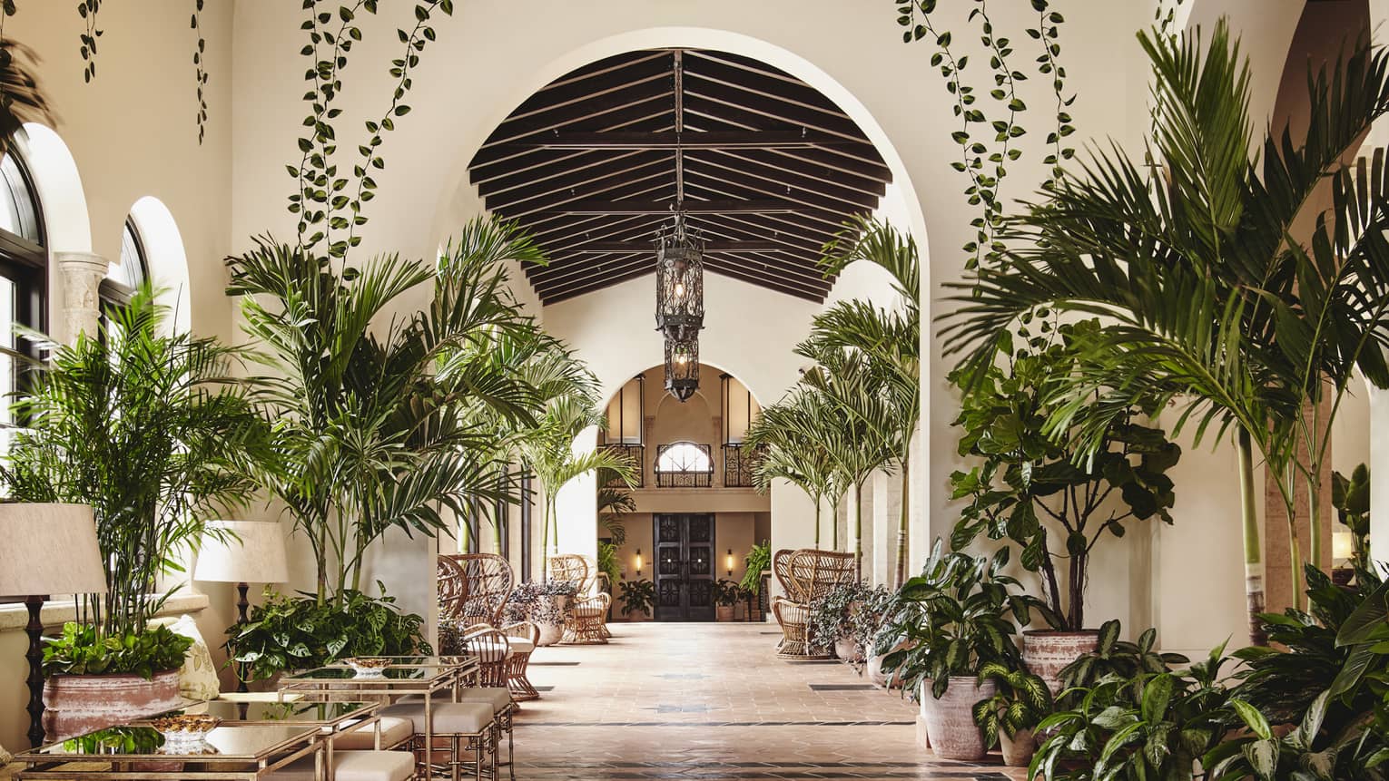 The interior of a lounge area of a hotel with small palm trees, other plants, and tables for people to sit at.