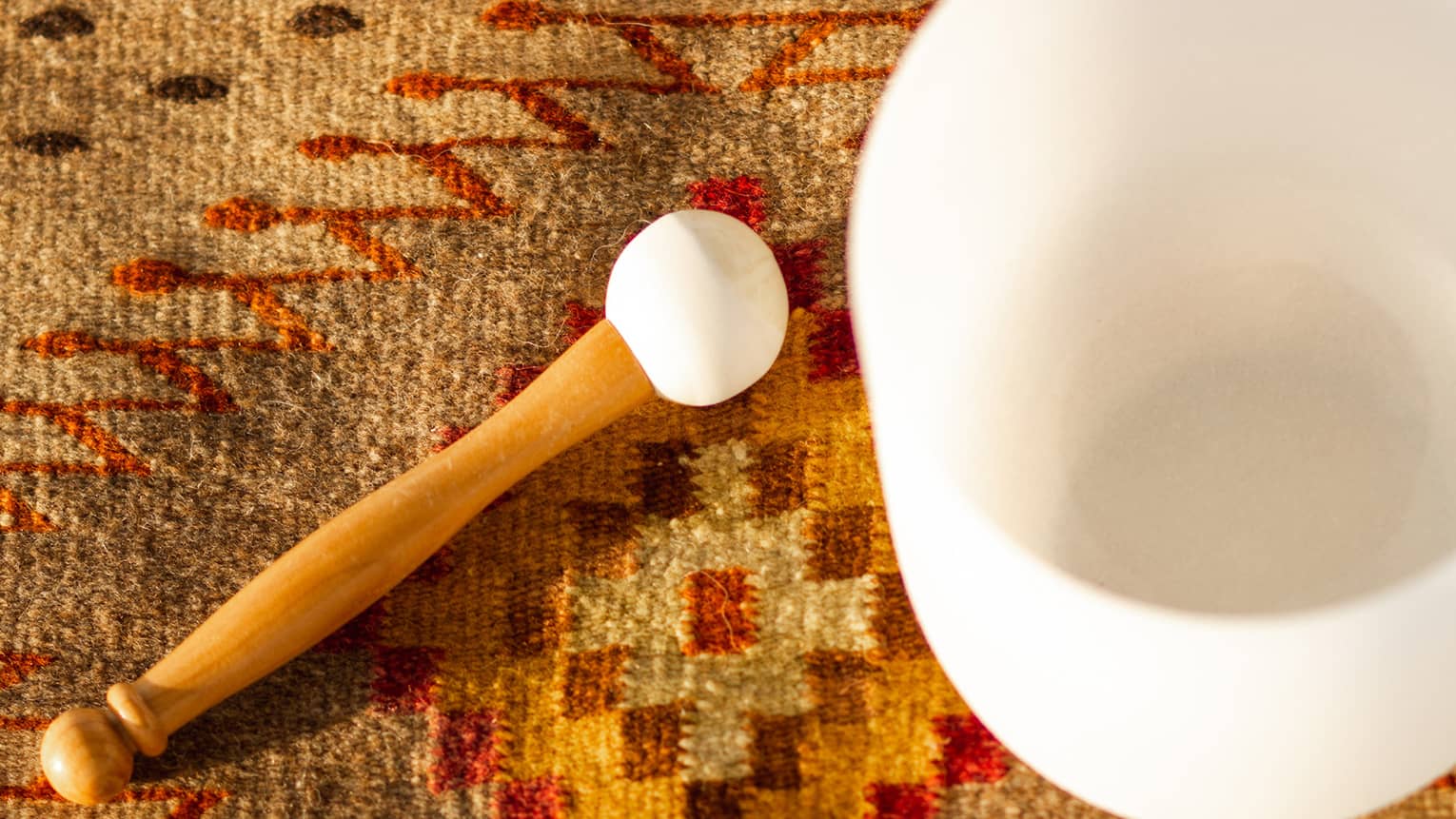 Wood mallet with white rubber tip rests on geometric Moroccan rug, beside a white crystal singing bowl.