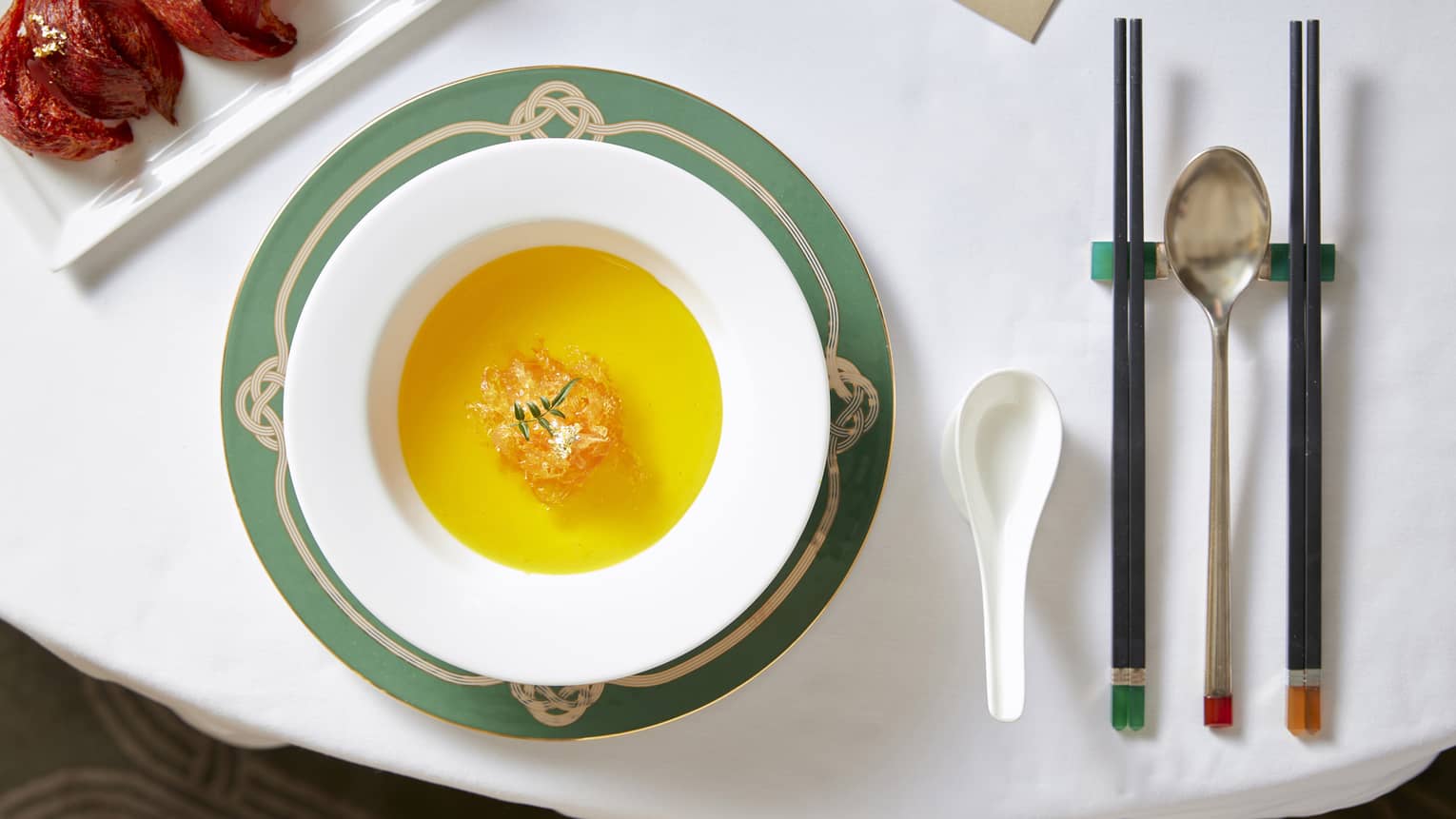 Golden bird’s nest soup in a white bowl on a decorative green and gold charger plate, set with chopsticks and spoons.