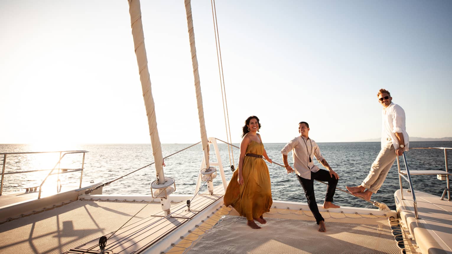 A woman wearing a flowing yellow dress, a man wearing a white shirt and black pants and another man wearing a white shirt and khaki pants stand on the back of a sailboat smiling and looking out onto the ocean