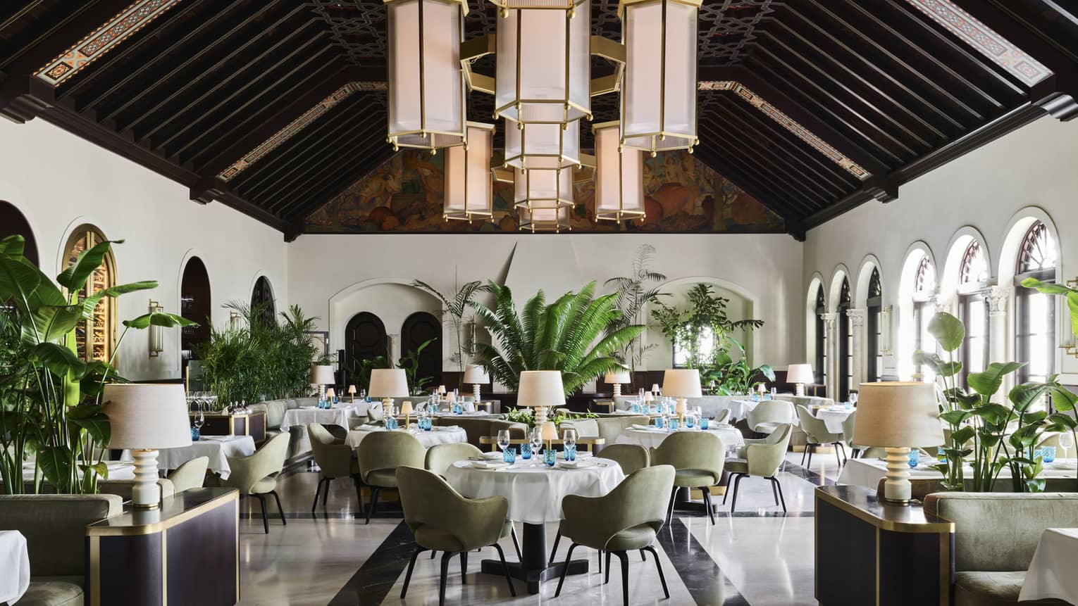 The light-filled Lido Restaurant with white walls, windows, dark wooden beams on a pitched ceiling and hanging lantern-style lights. One of the many Surfside restaurants of note at Four Seasons Surf Club, Florida.