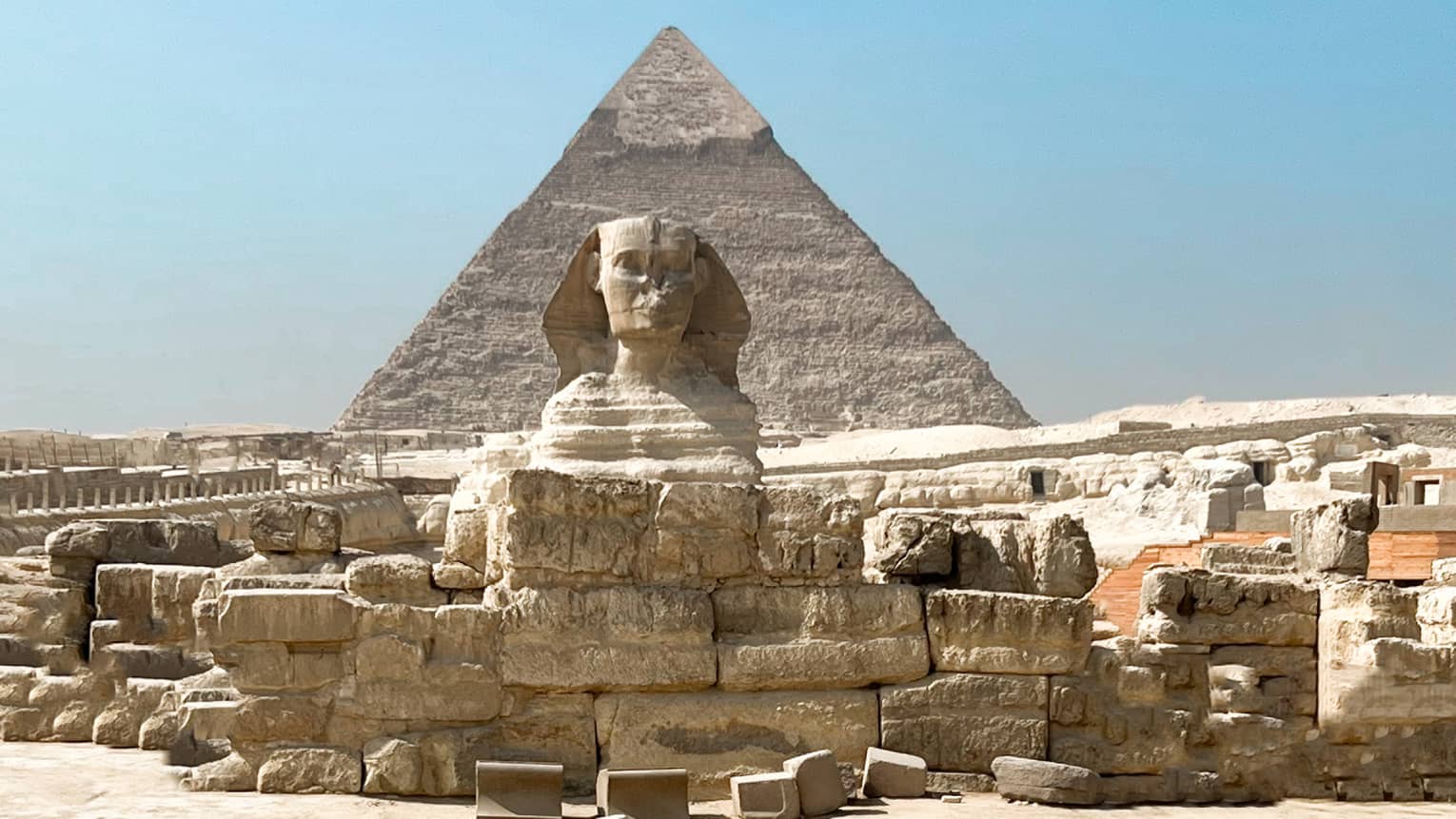 View from behind as a guest crosses golden desert sand toward the immense ruins and sphinx, a pyramid rising up beyond.