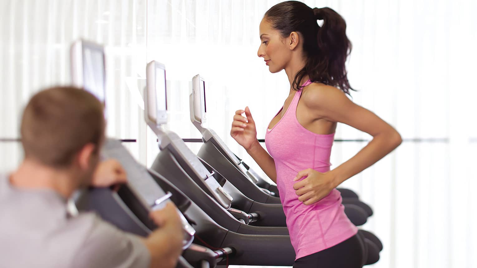Woman in pink sleeveless shirt and ponytail runs on treadmill as personal trainer watches