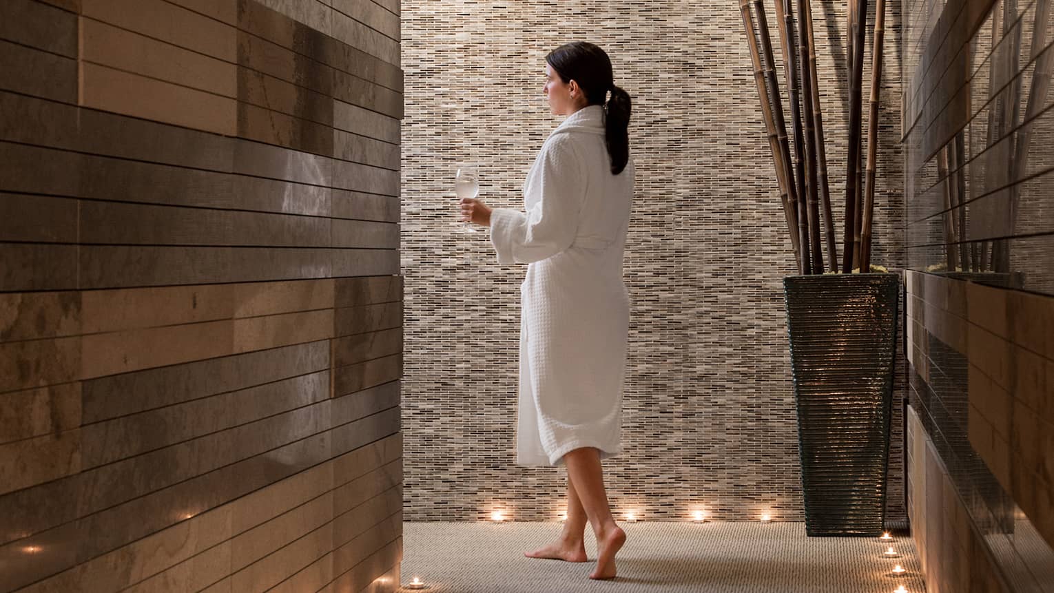 Woman in bathrobe holding wine glass with water walks through spa