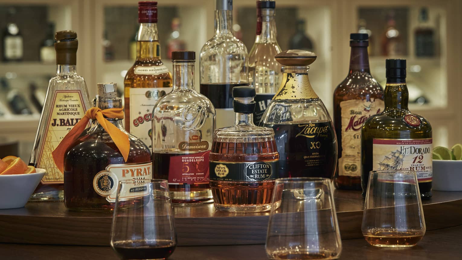 A rum tasting set-up on a brown restaurant table with a variety of glass rum bottles in the centre, small bowls of citrus slices to either side and three tasting glasses of rum in front