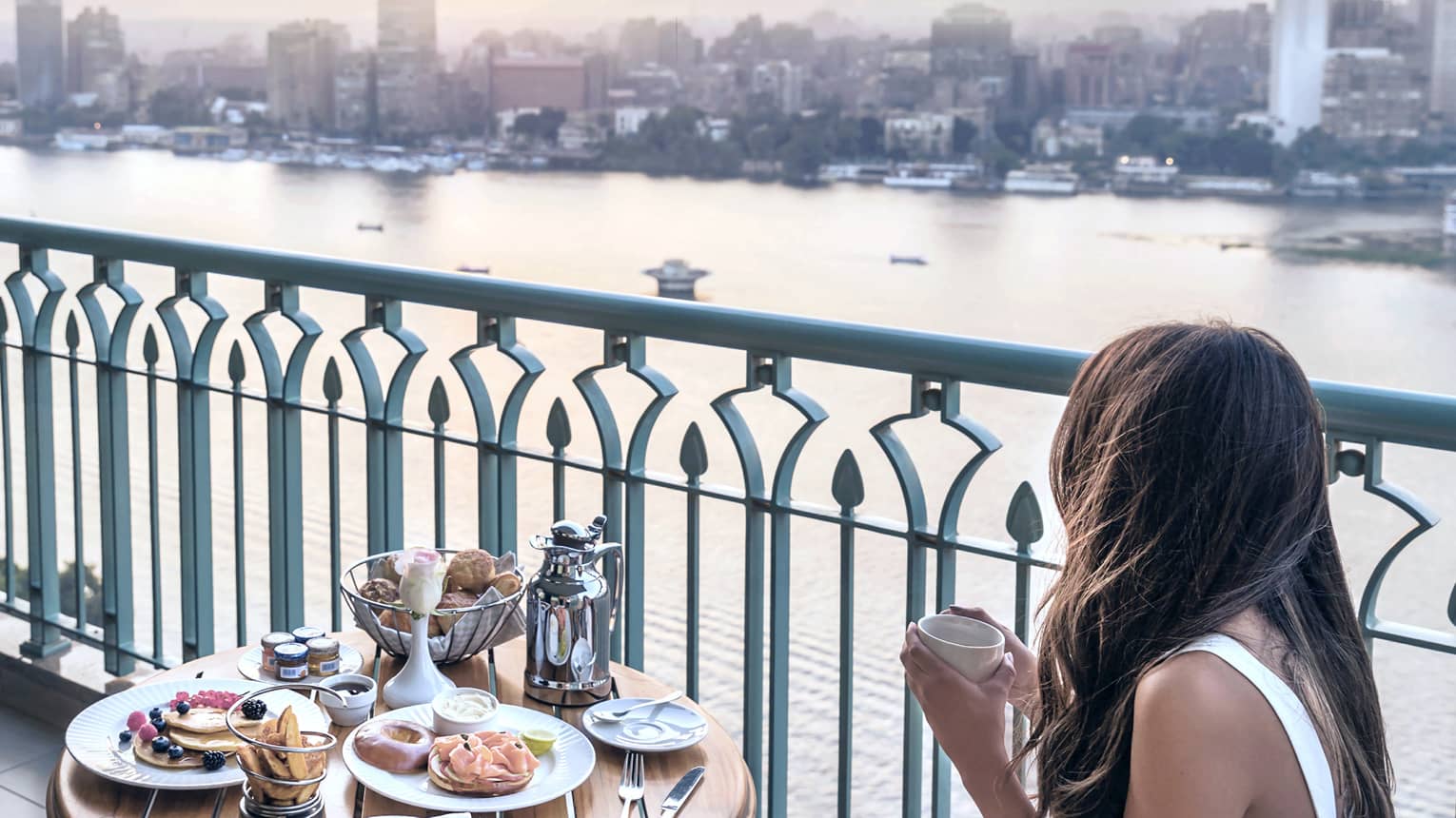 Dark-haired woman in white dress seated at balcony bistro table looking through railing at river and city views