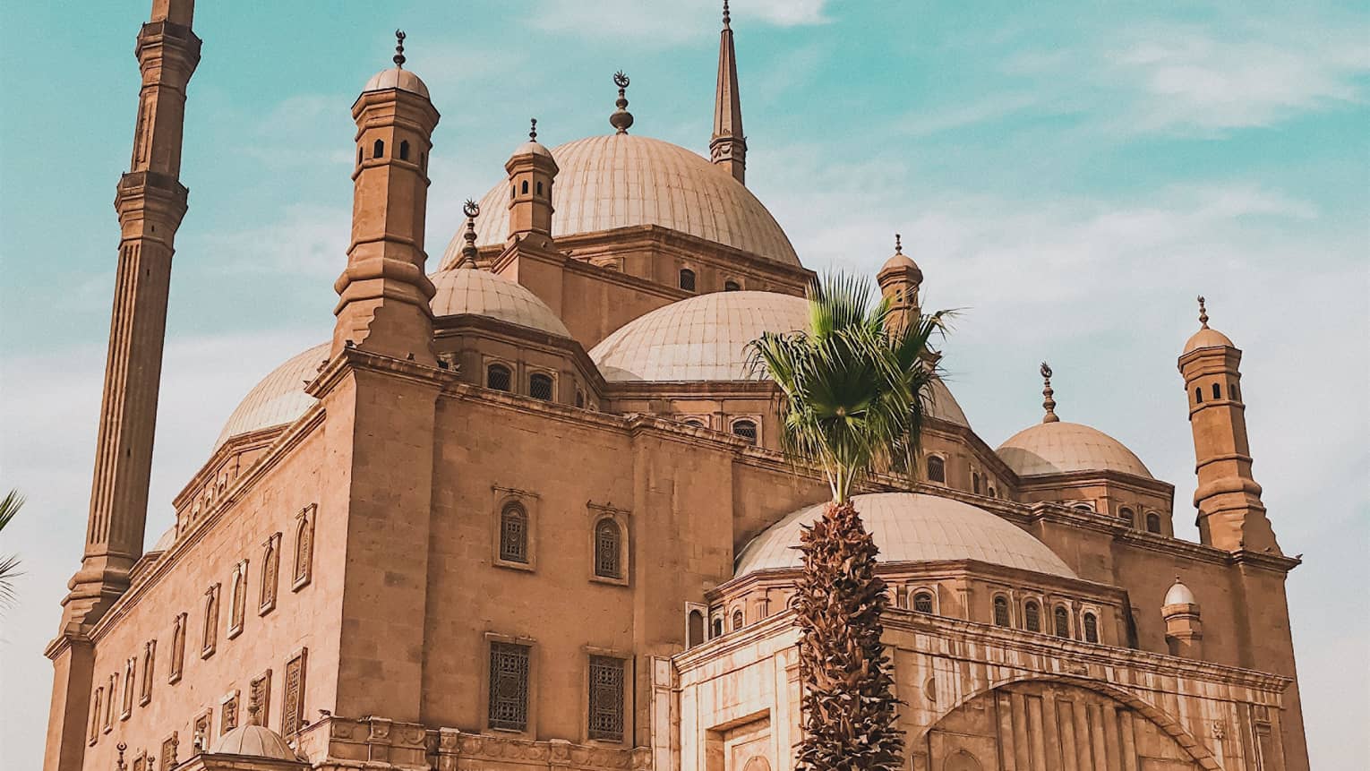 A tourist poses in front of the imposing Mosque of Muhammad Ali, minarets towering above the many domes of the building.