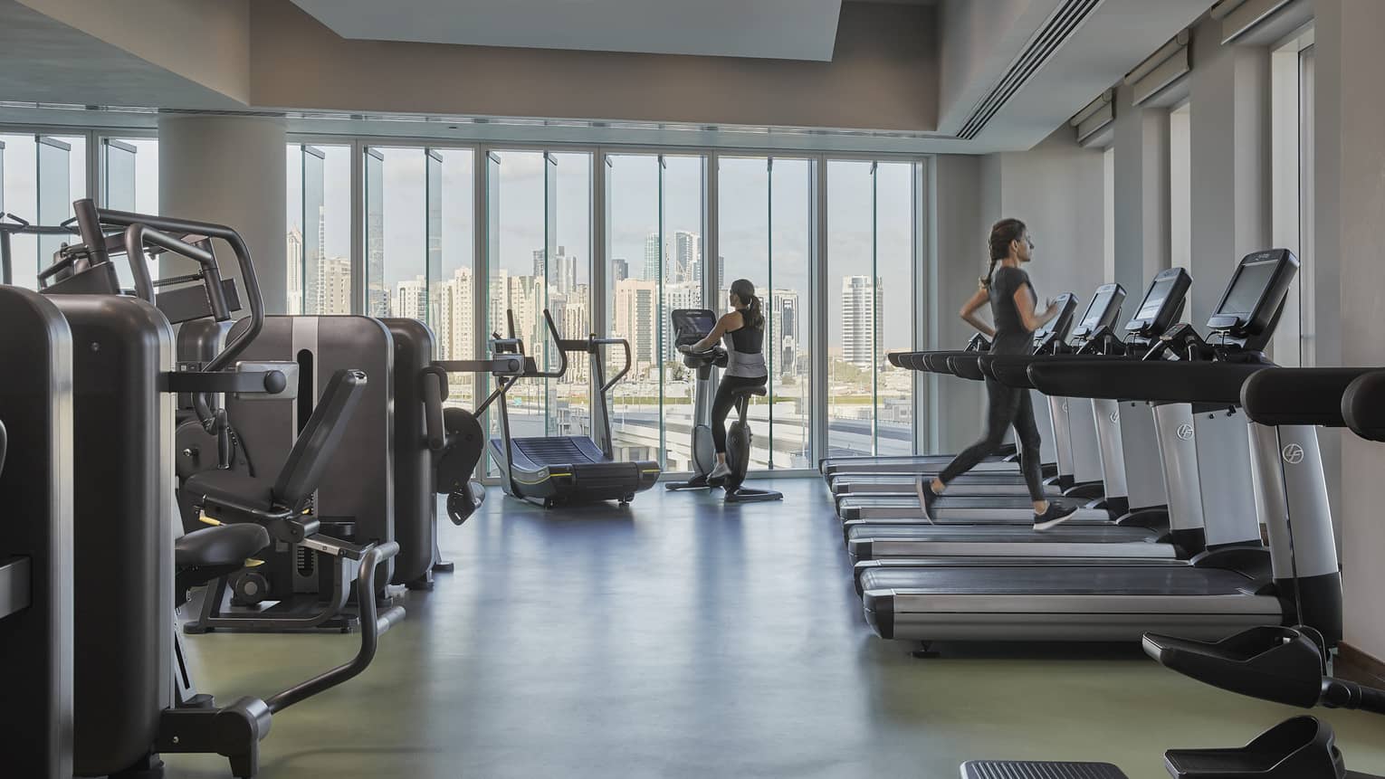 Two women working out on a treadmill and an eliptical in a large grey gym overlooking the Abu Dhabi skyline.