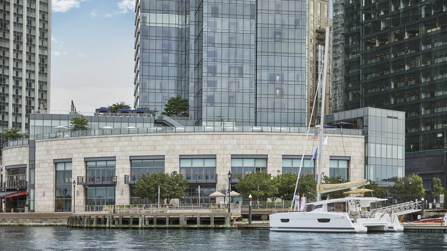 A sail boat on the water next to a large brick building and a pier, with tall skyscrapers in the background.