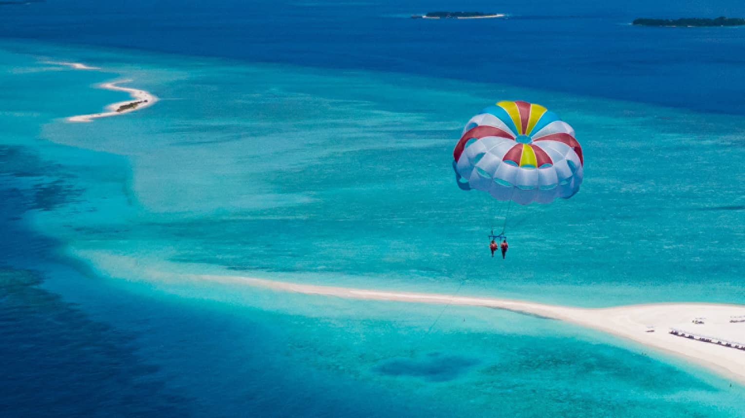Aerial view of two people parasailing in tandem, colourful sail floating above an expanse of aqua water and a white sandbar.