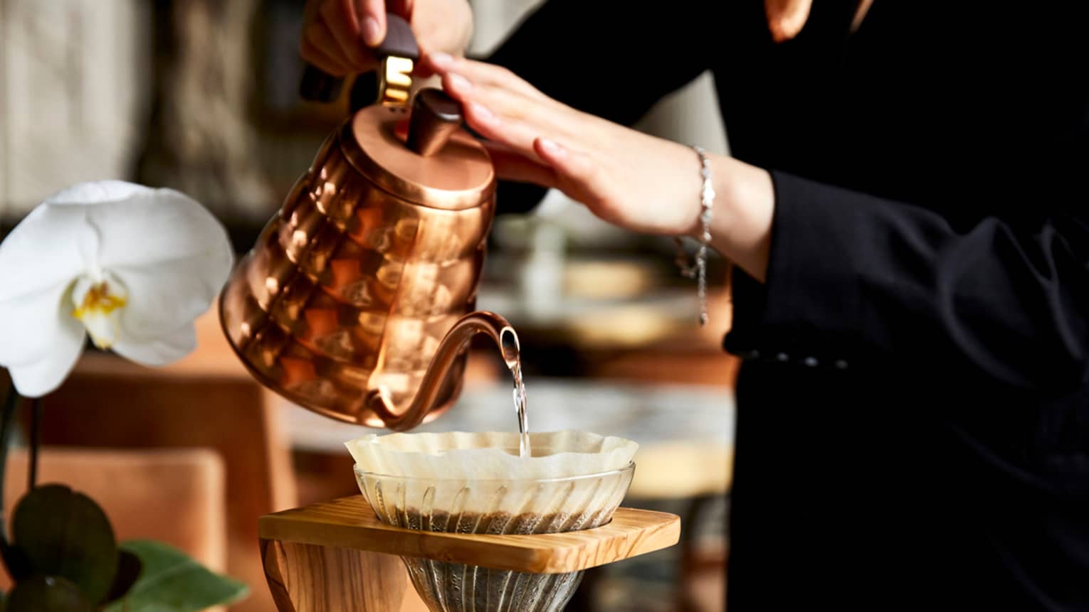 A woman pours water into a filter filled with coffee grounds, which drips into a glass container of hot coffee